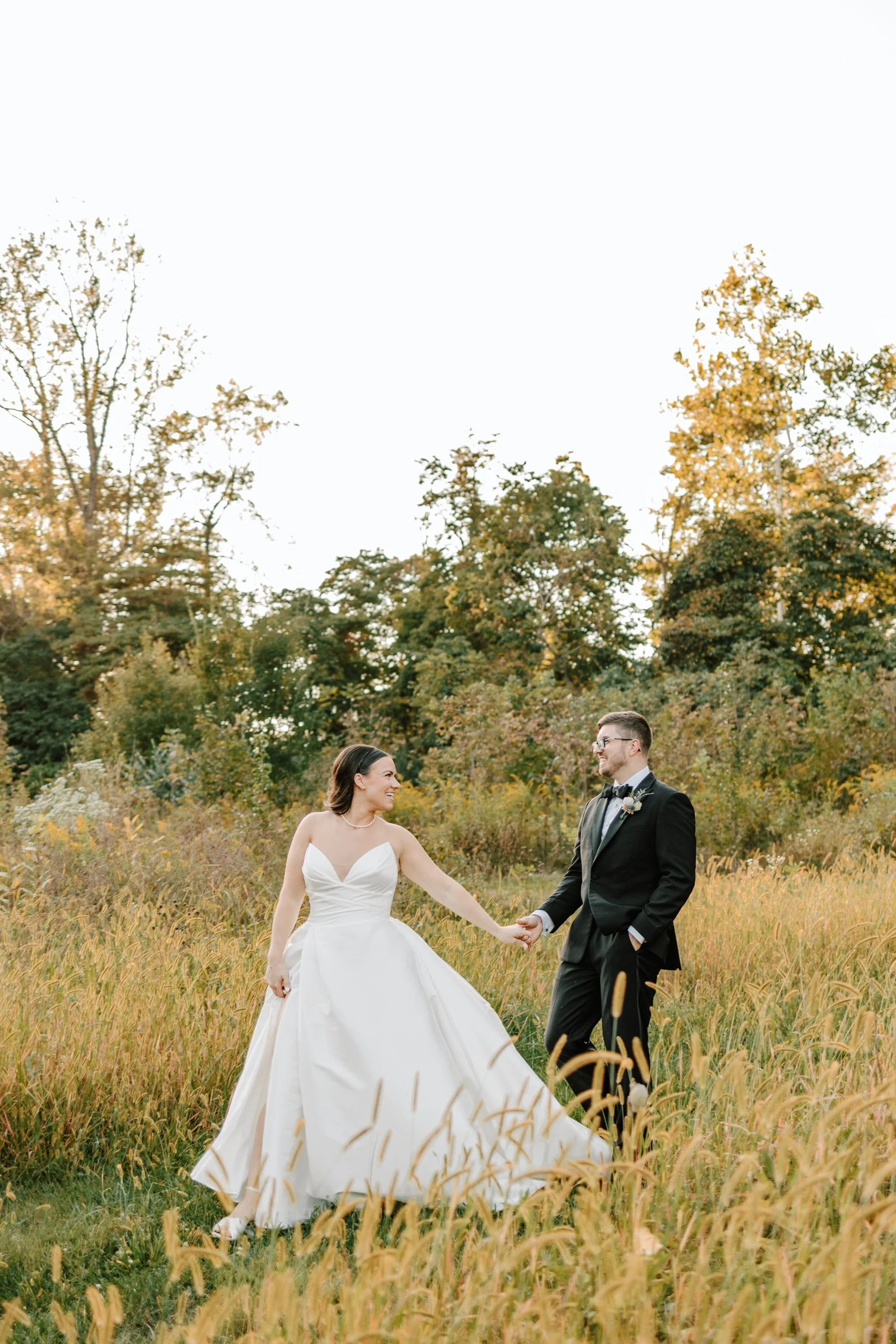 A bride and groom holding hands in a field with tall grass, trees in the background, during sunset.