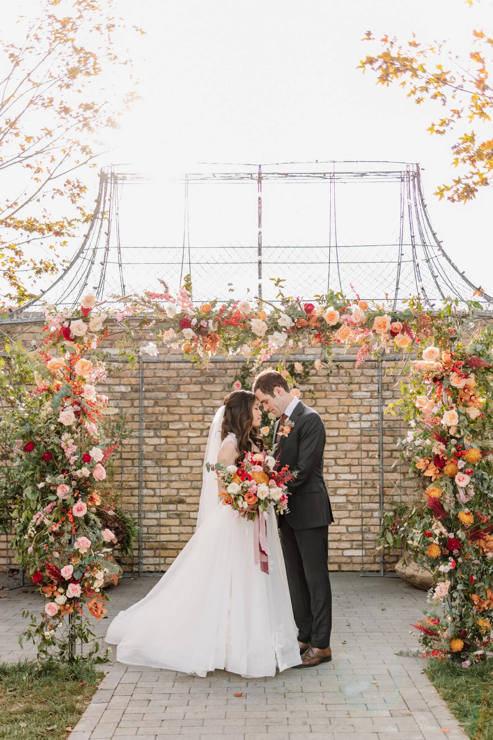A bride and groom stand under a floral archway during their wedding ceremony, embracing with foreheads touching in front of a brick wall and an outdoor sky.