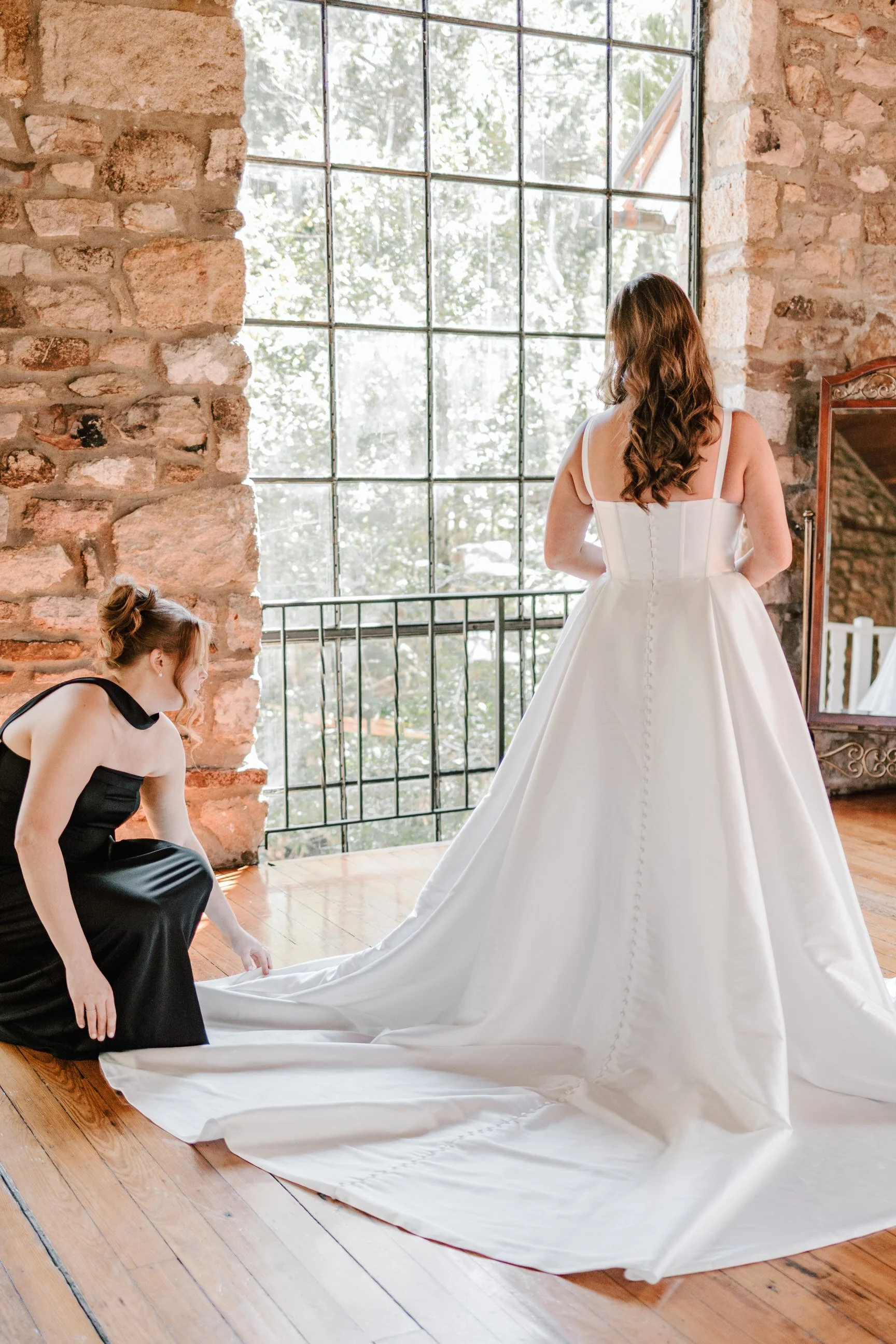 A woman in a wedding dress is being assisted by another woman in a black dress to prepare for her wedding. They are in a room with large windows and exposed brick walls.