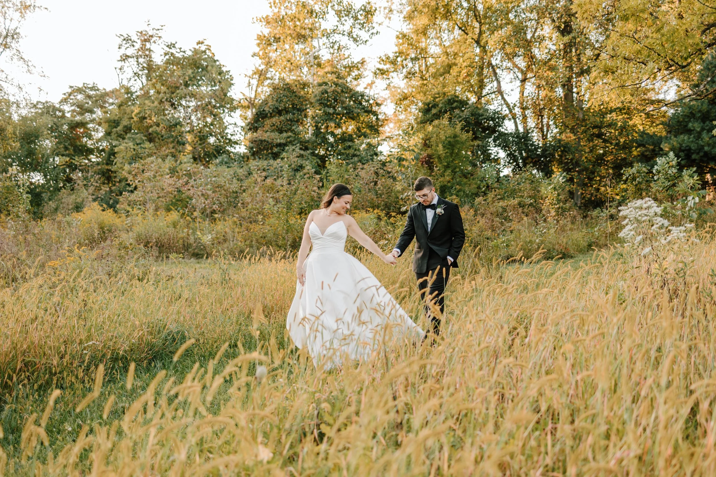 A bride and groom walking hand in hand through a grassy field during autumn, surrounded by trees with yellow and green leaves.