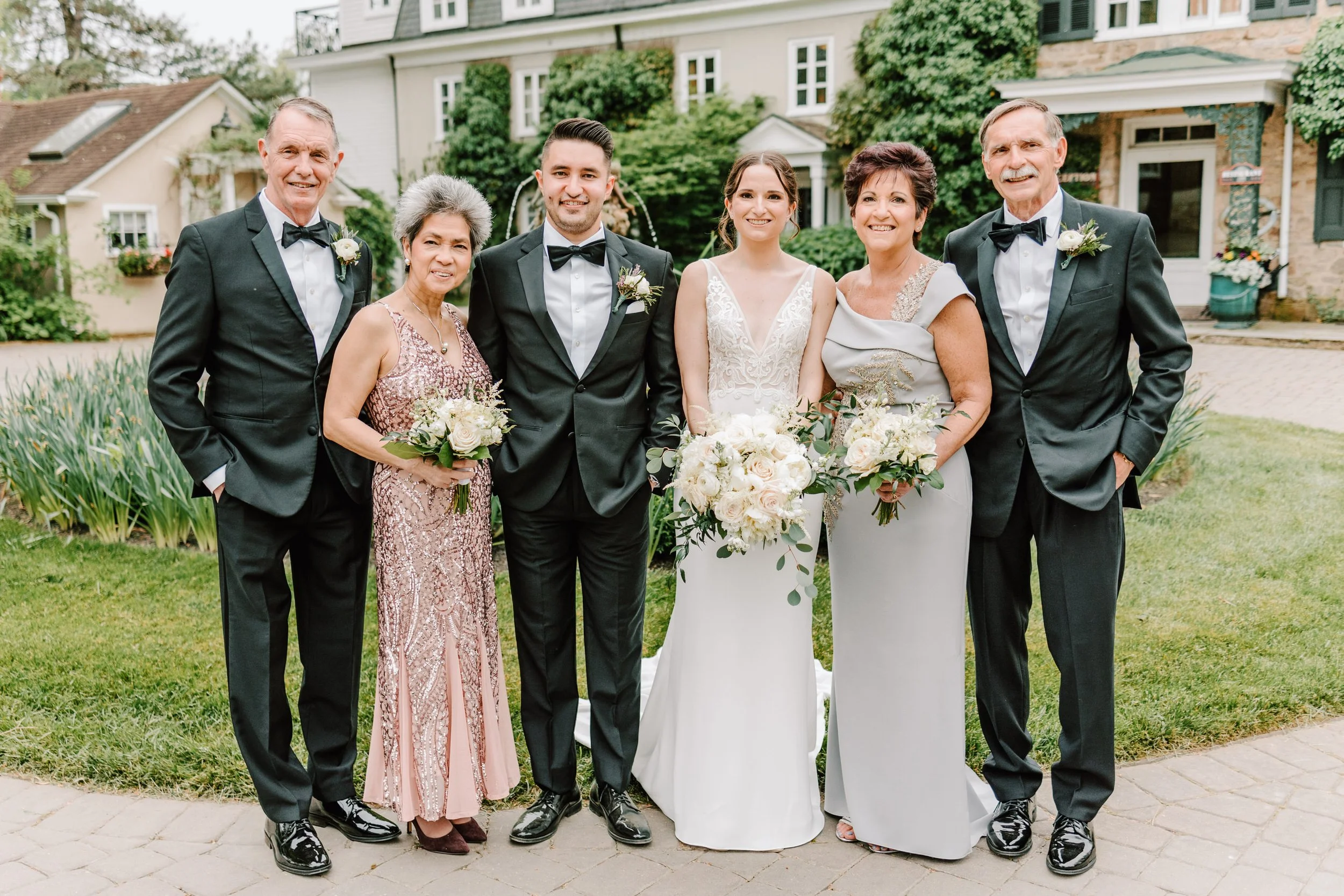Group of seven people, including bride and groom, standing outside in front of a house with greenery. The bride wears a white wedding dress and holds a bouquet. The groom wears a black tuxedo. The other five people, two men and three women, are dress