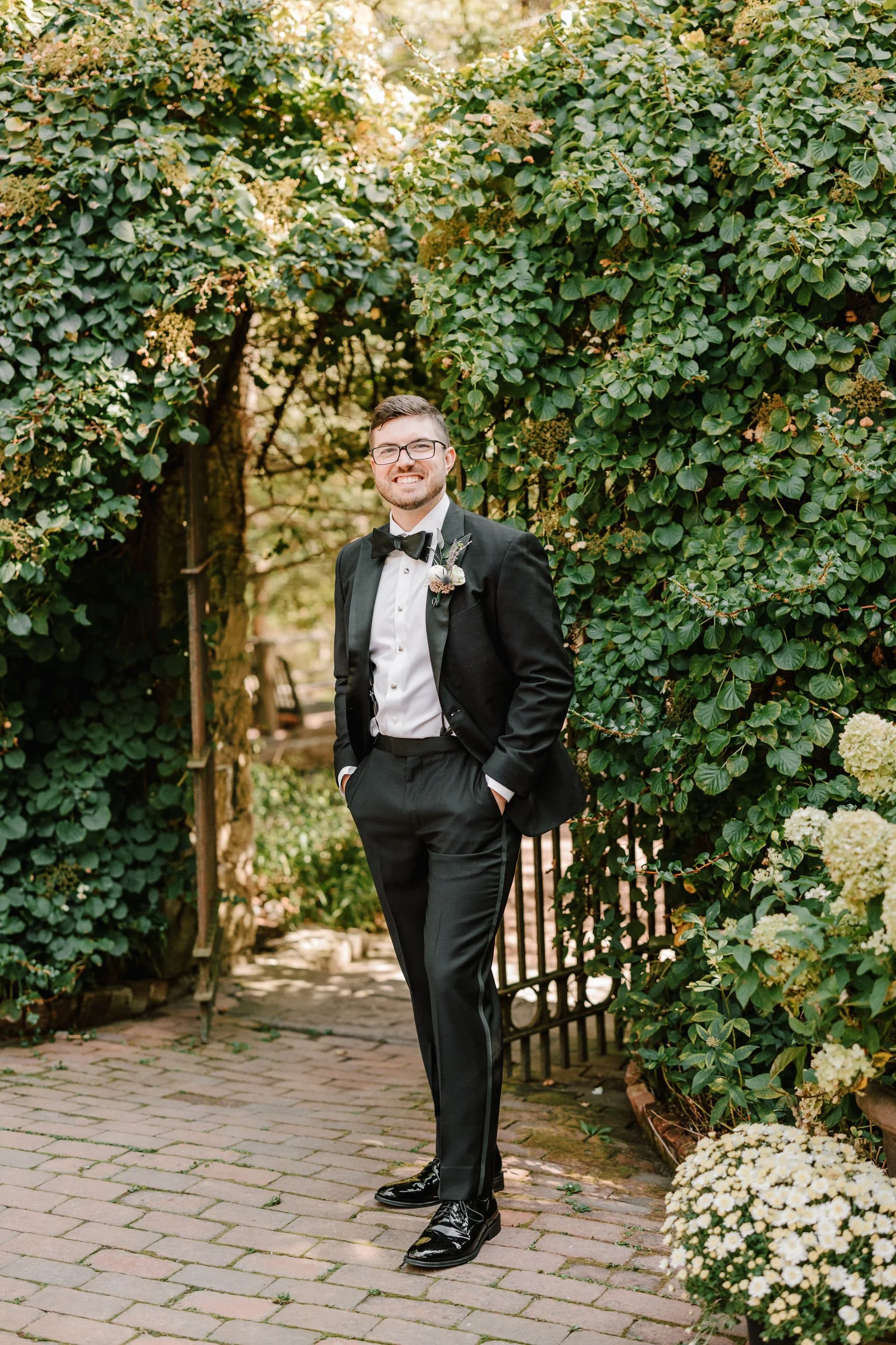 A man in a tuxedo stands outdoors among green foliage and white flowers, smiling at the camera with hands in pockets.