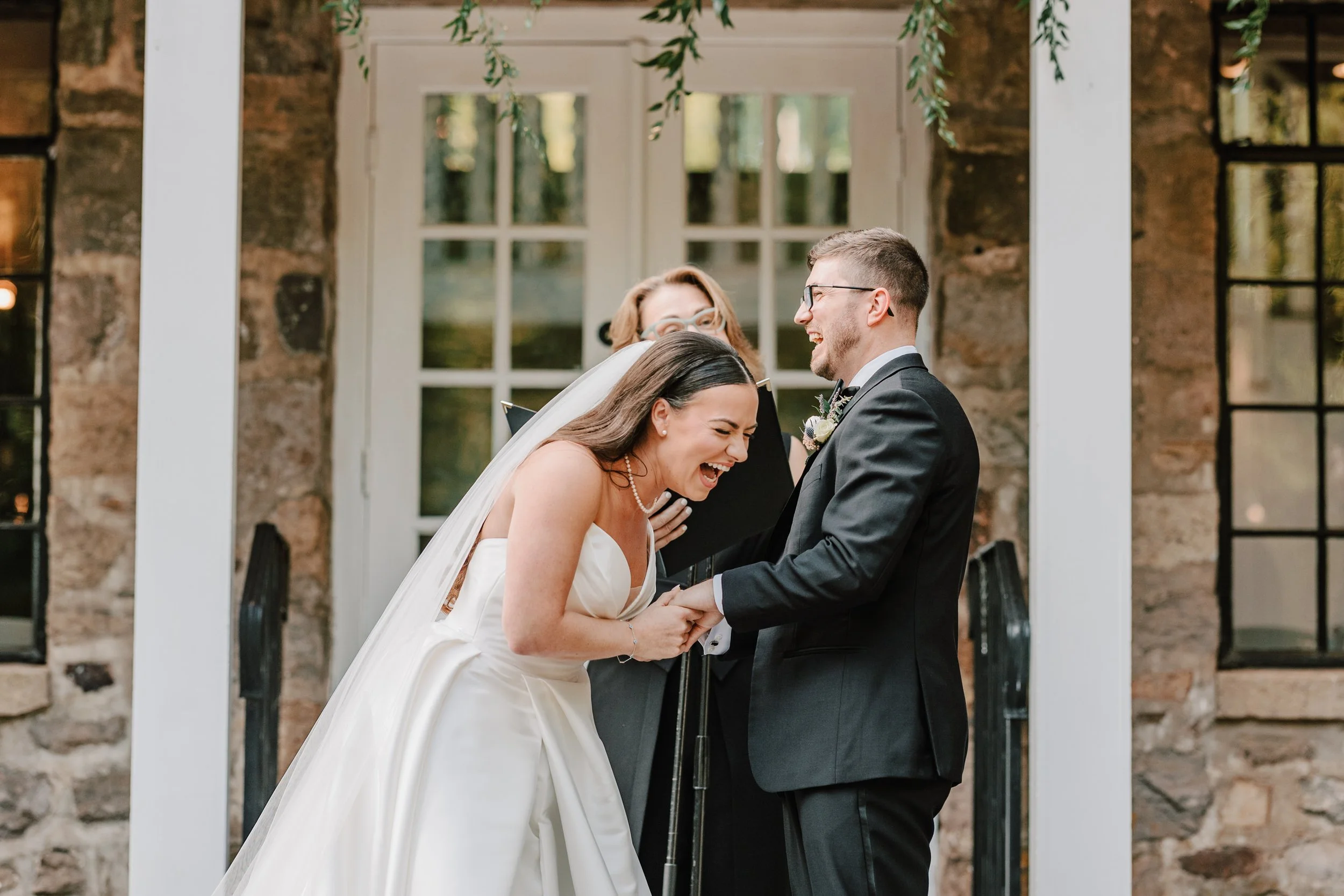 A bride and groom laughing and holding hands during their wedding ceremony outside a stone building with large windows, as an officiant stands behind them.