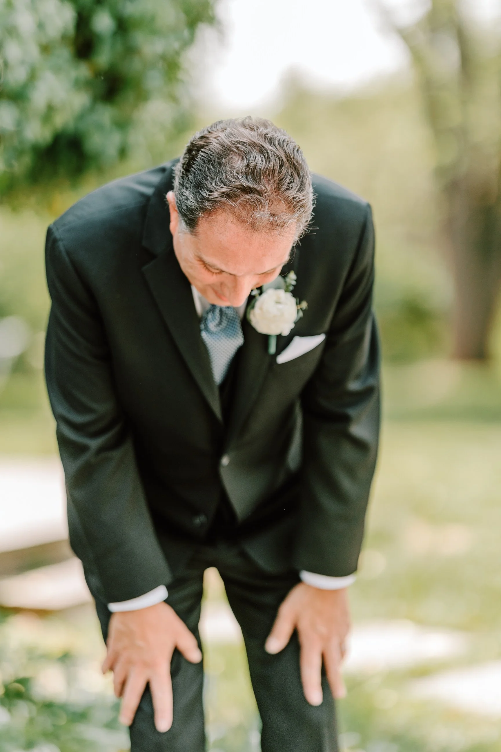 An elderly man in a black tuxedo, white shirt, and bow tie, with a white boutonniere, bows his head while outdoors.