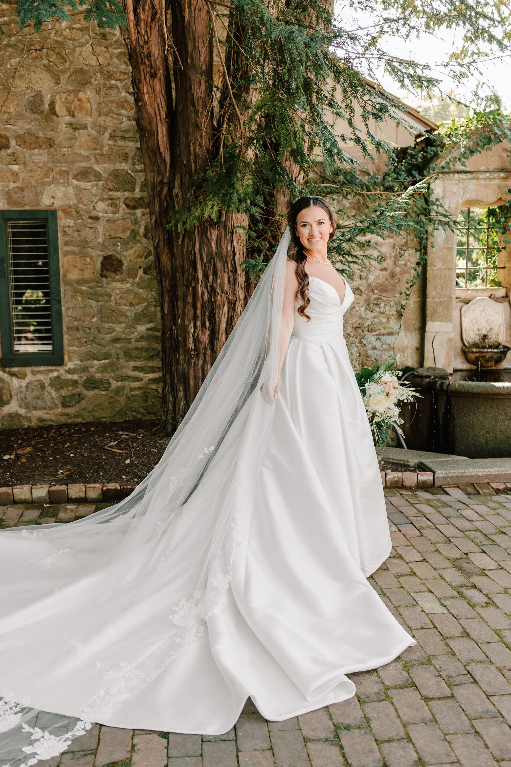 A bride in a white wedding gown standing outdoors on a brick pathway, with a large tree and an old stone building in the background.