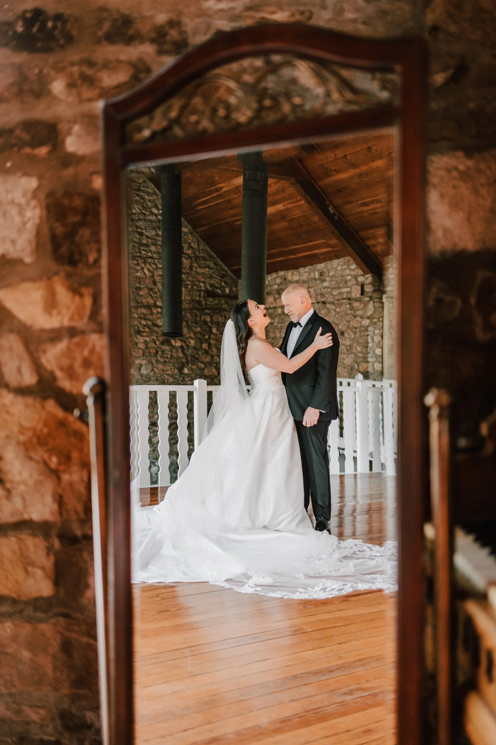 A bride and groom dancing together on a wooden floor, reflected in a mirror, with stone walls and wooden ceiling in the background.