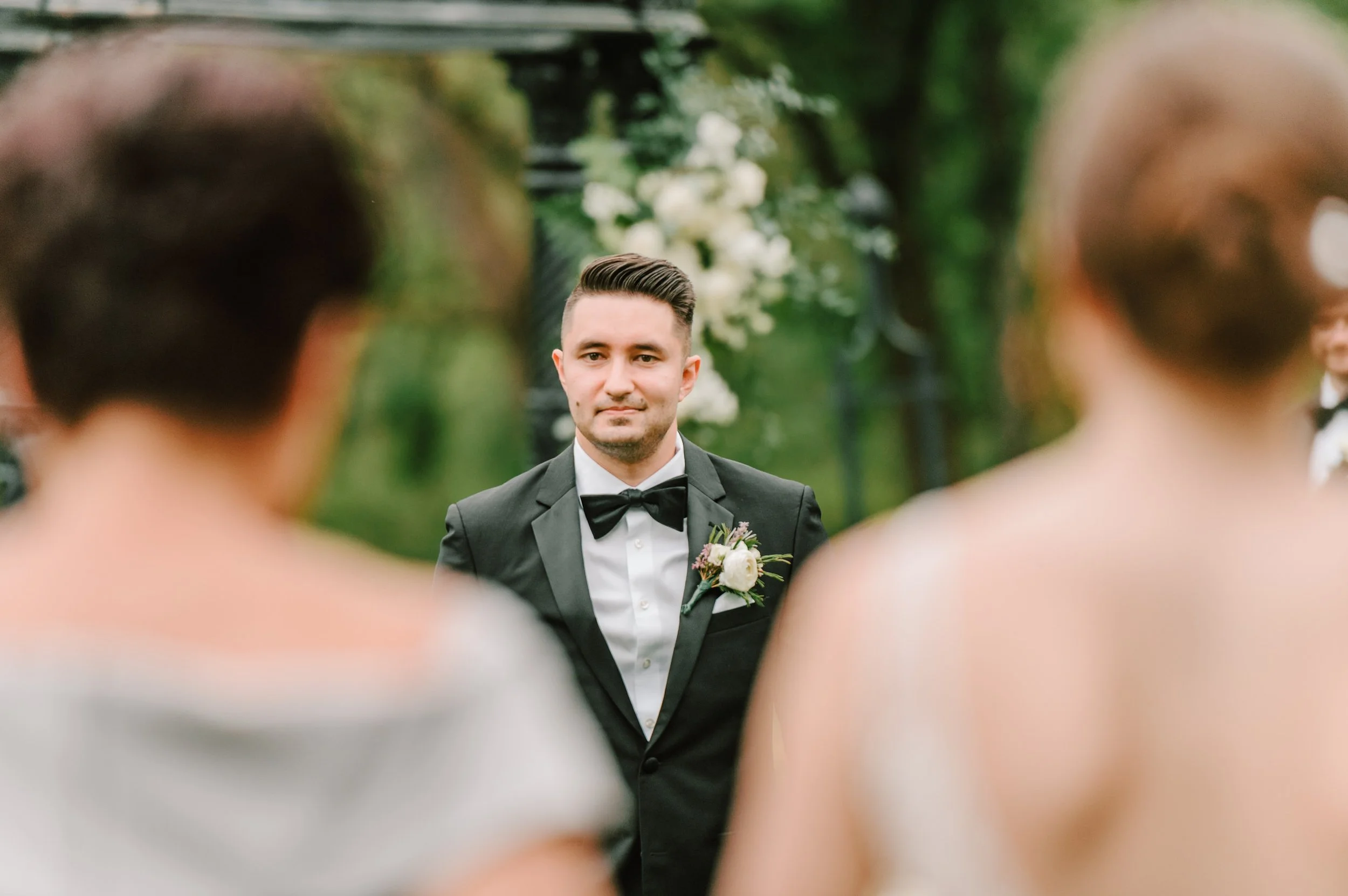A groom in a black tuxedo and bow tie stands outdoors during a wedding ceremony, surrounded by blurred guests, with greenery and white flowers in the background.