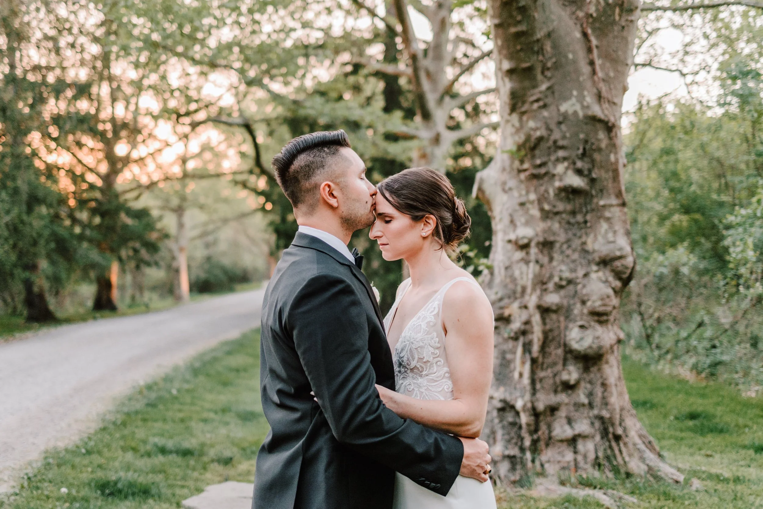 A newlywed couple standing outdoors with the groom kissing the bride on the forehead. The bride has closed eyes, wearing a white dress with lace detail, and the groom is in a black tuxedo. They are next to a large tree with a park path in the backgro