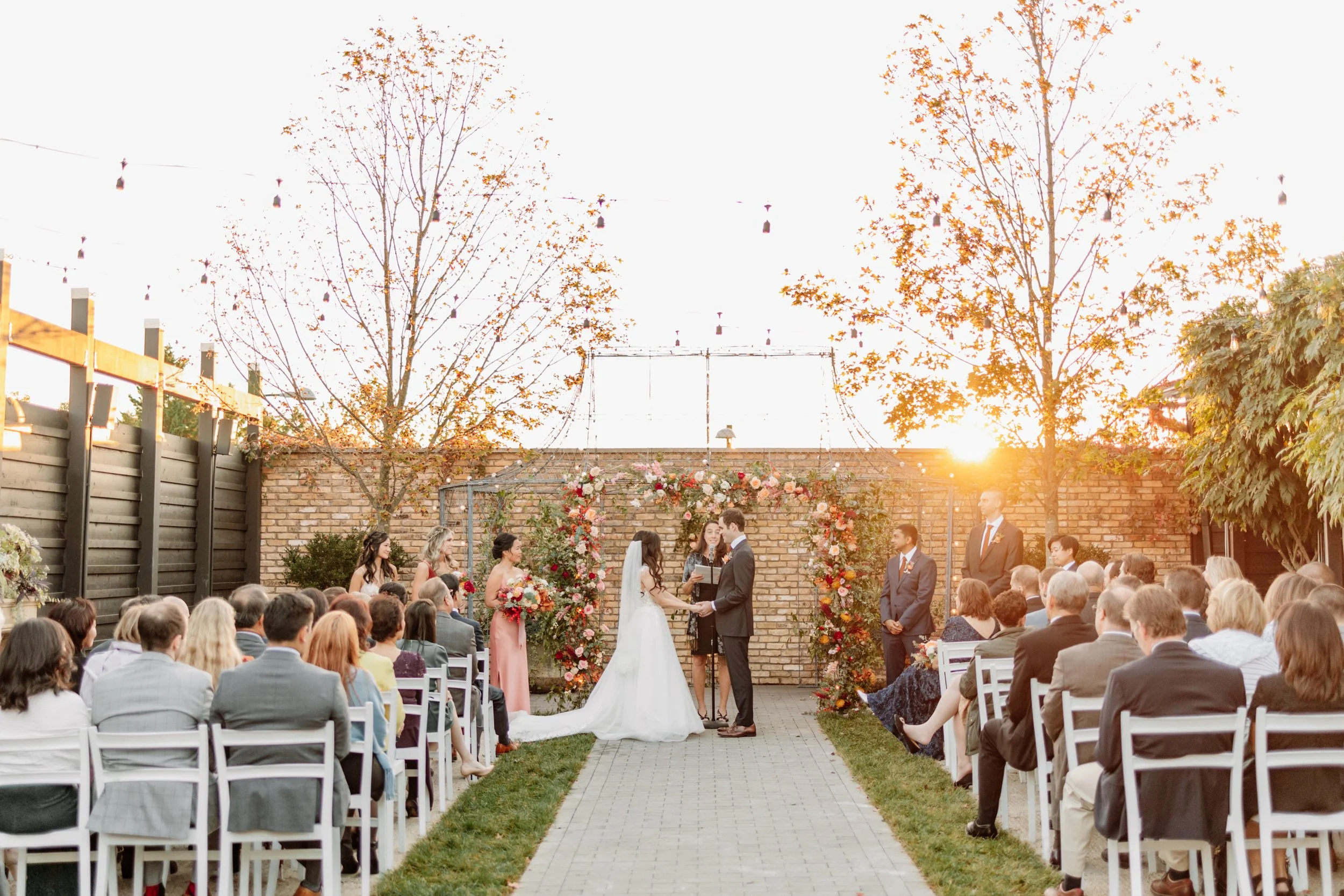 A backyard wedding ceremony at sunset with a couple exchanging vows, surrounded by guests seated on white chairs, floral decorations, and two trees.