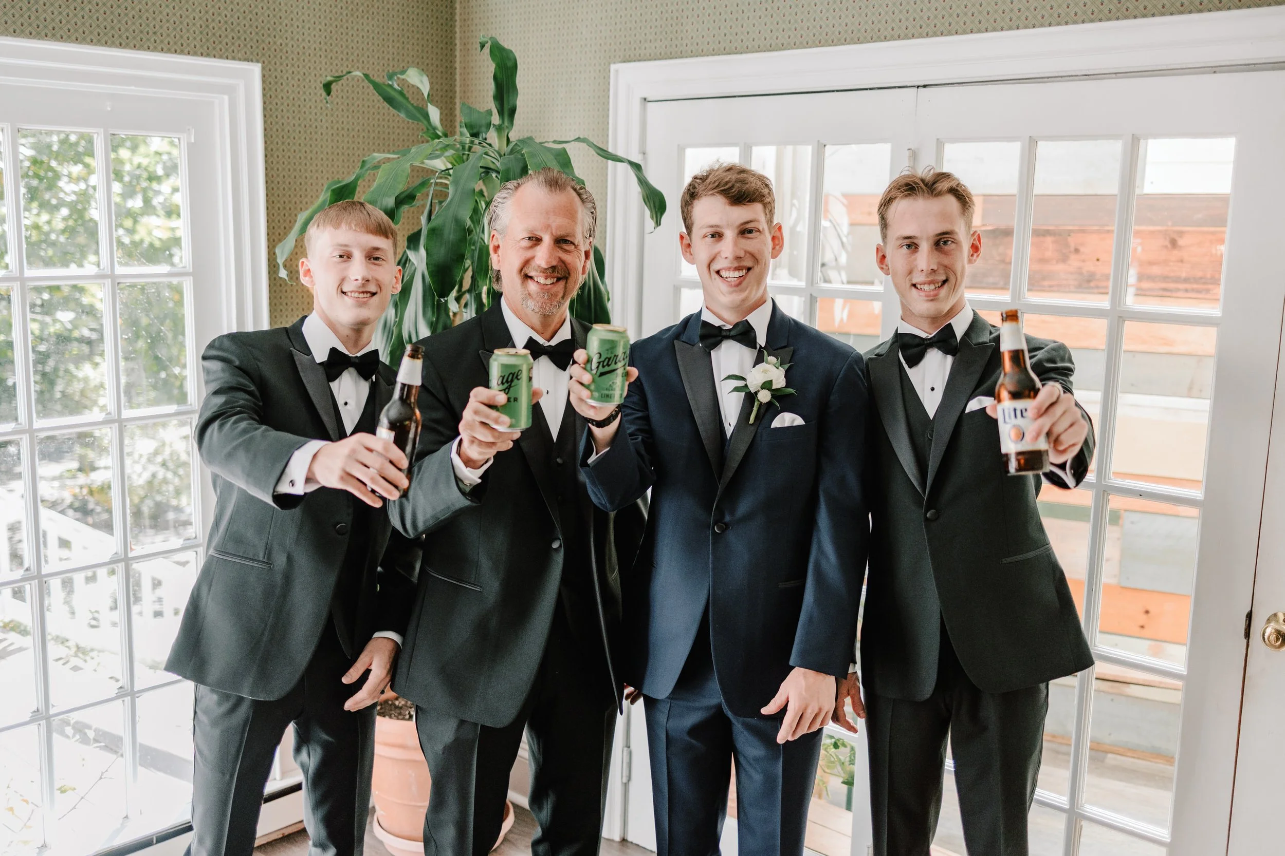 Four men in tuxedos celebrating with drinks indoors, standing in front of a glass door, smiling at the camera.
