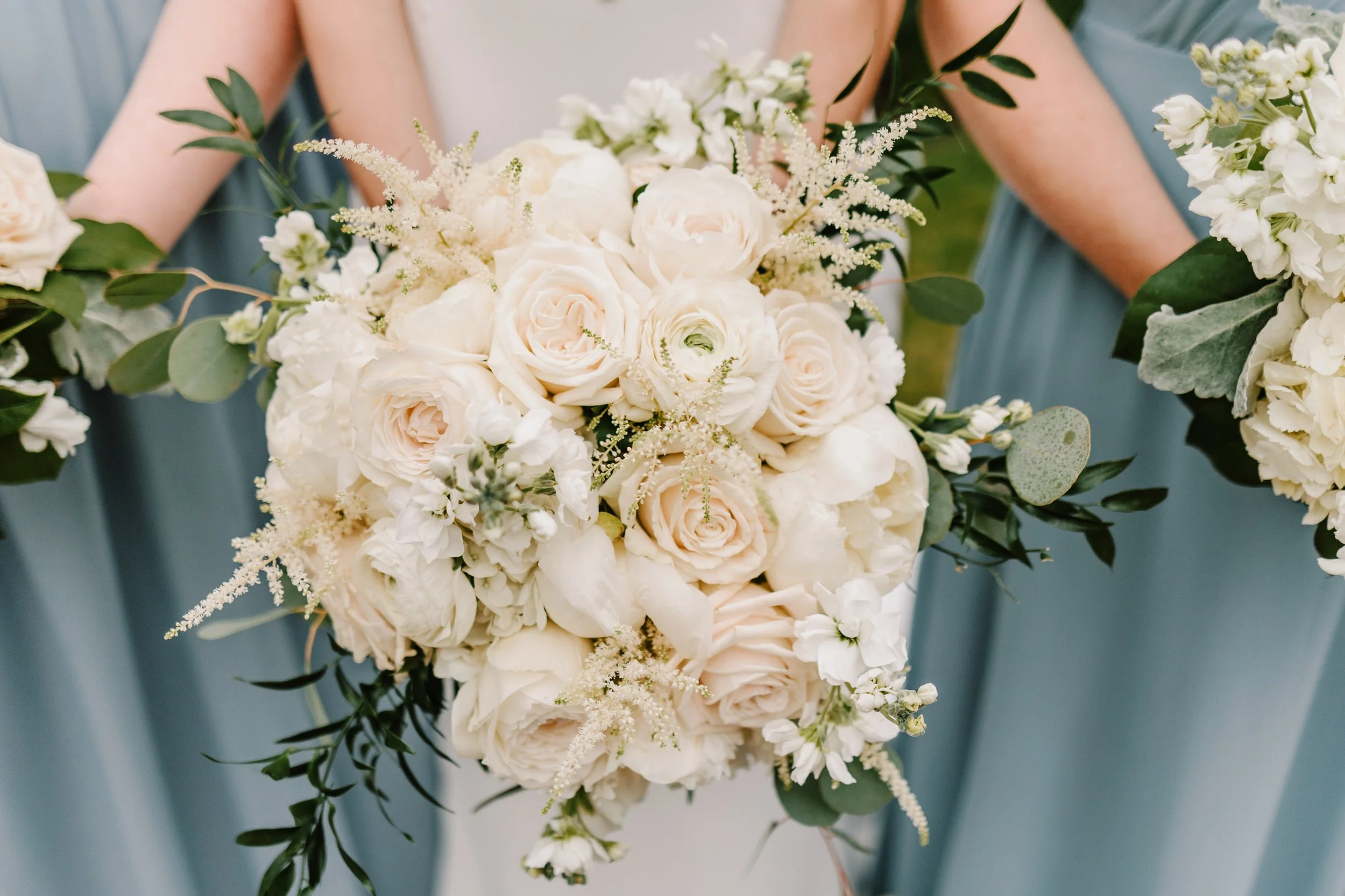 Bridal bouquet with white roses, peonies, and greenery held by a bride wearing a light blue dress at a wedding.