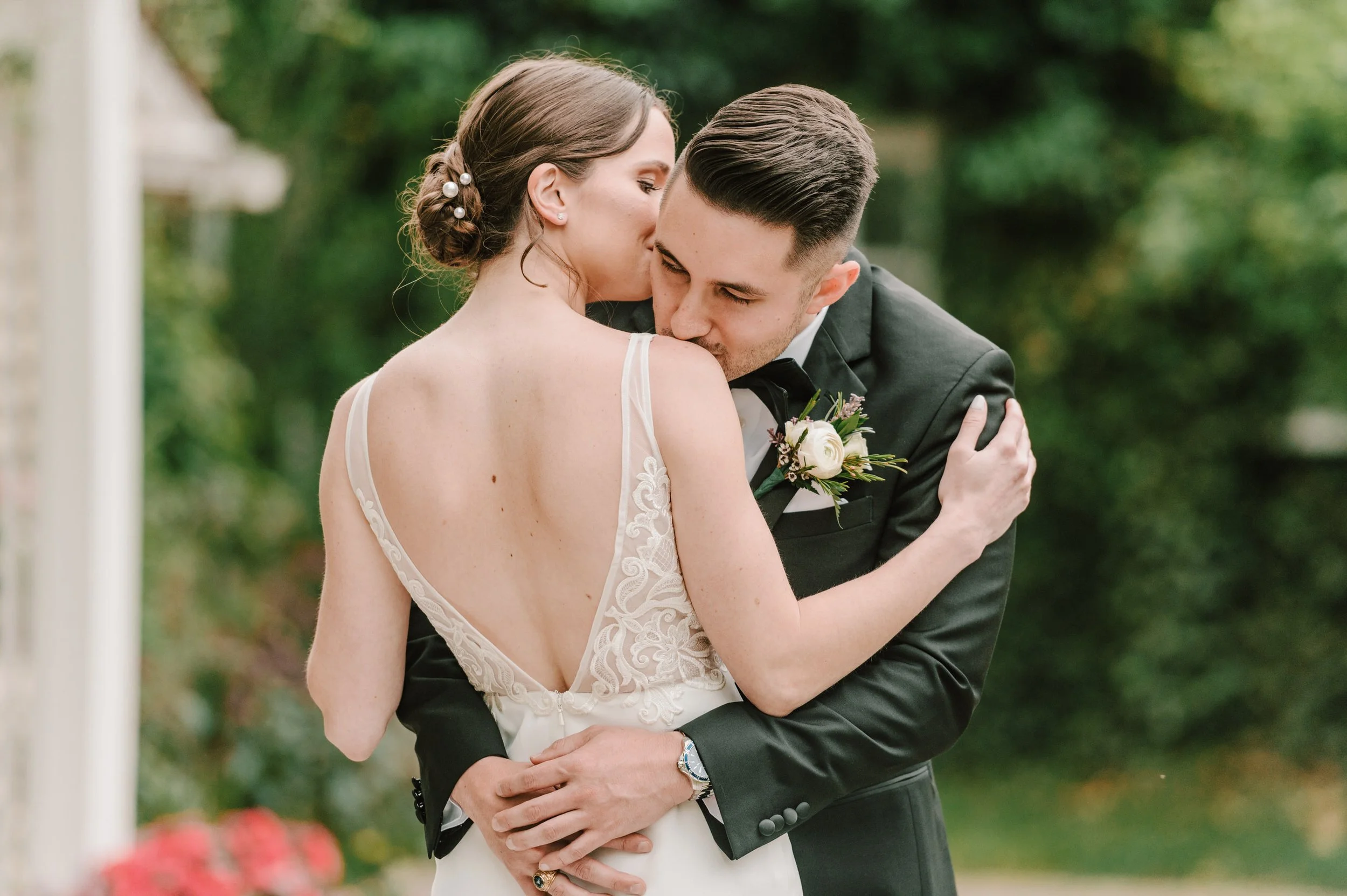 A bride and groom embracing, with the bride whispering into the groom's ear or kissing him on the cheek, outdoors with greenery in the background.
