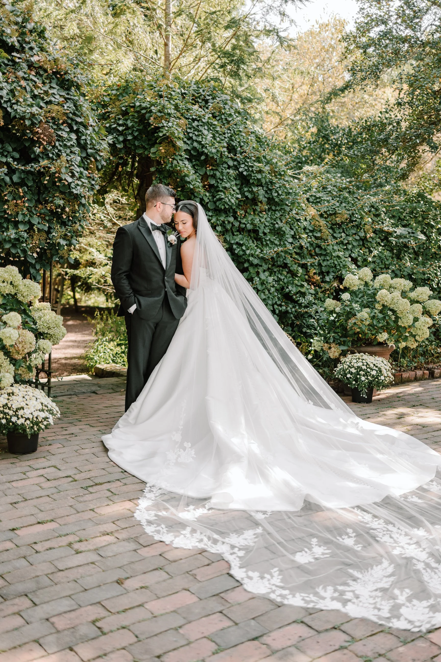A bride and groom standing close together outdoors, surrounded by lush greenery and flowering plants, with the bride wearing a white wedding gown and veil, and the groom in a black tuxedo.
