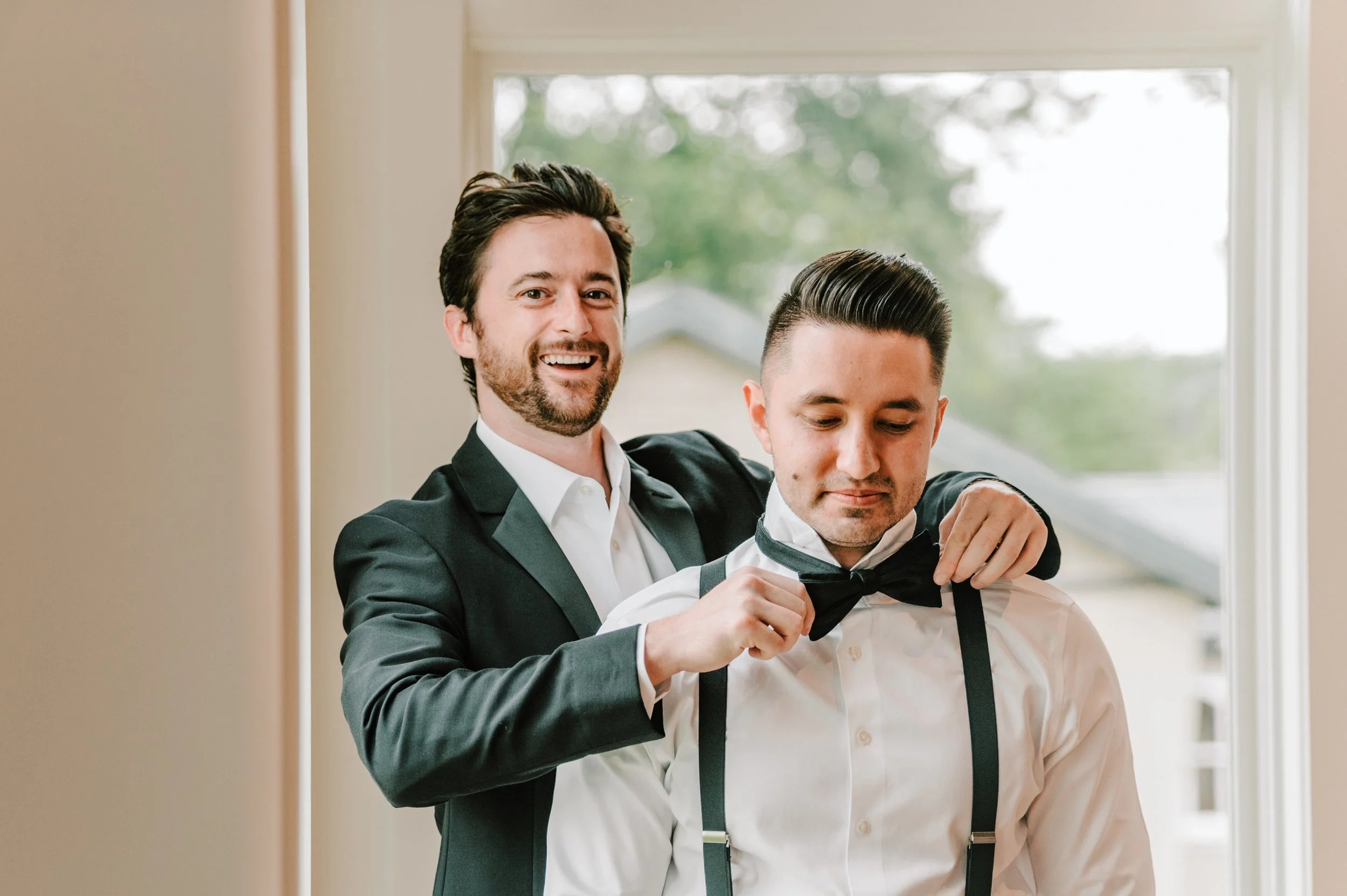 Two men, one smiling, helping the other with a bow tie in a room with a window behind them.