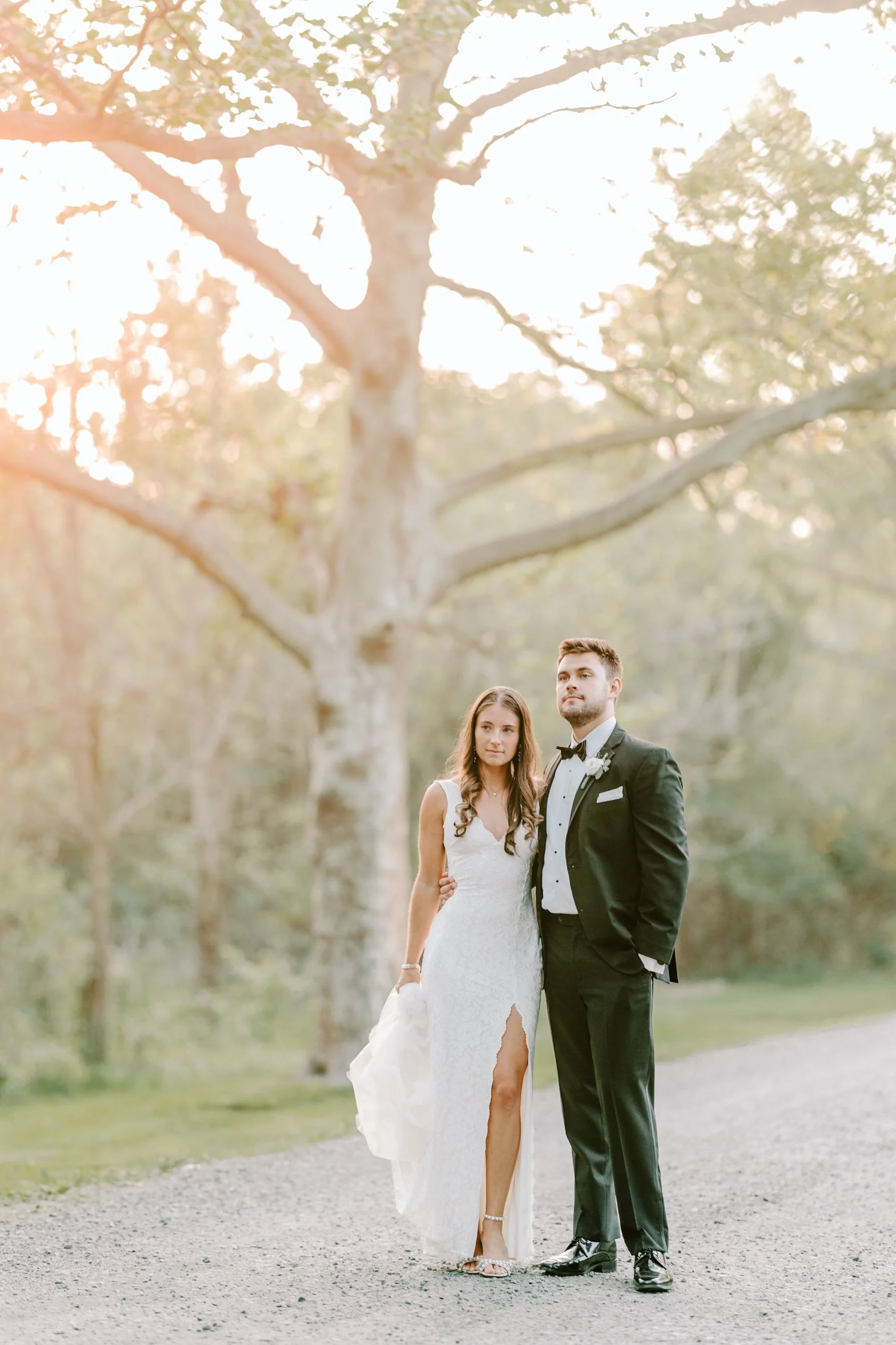 A bride and groom standing on a gravel path outdoors in front of a large tree during sunset, with the bride in a white dress and the groom in a black tuxedo.