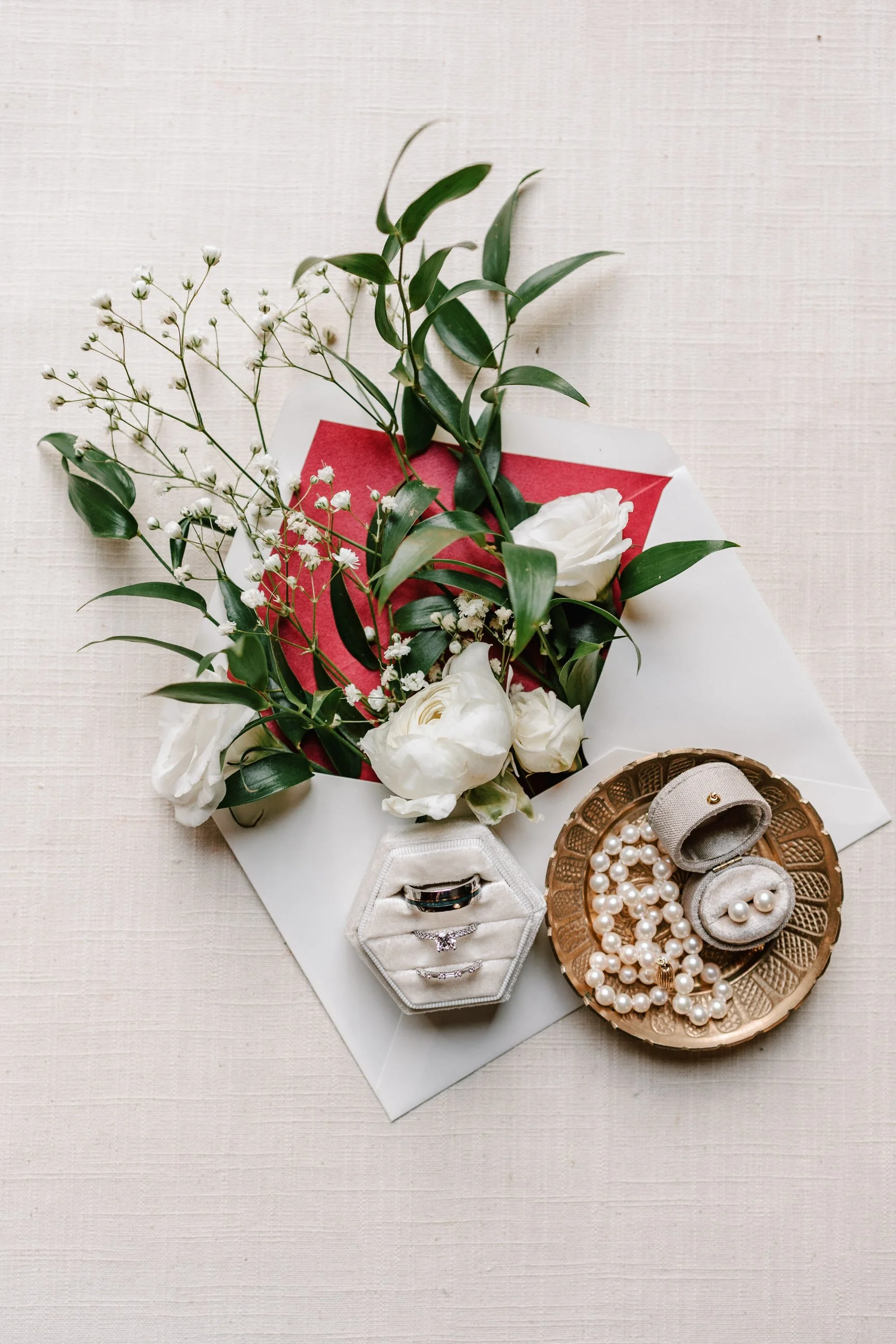 A bouquet of white roses and greenery, jewelry including pearl necklaces, bracelets, and rings displayed on a gold tray and in velvet jewelry boxes, all arranged on a beige fabric surface.