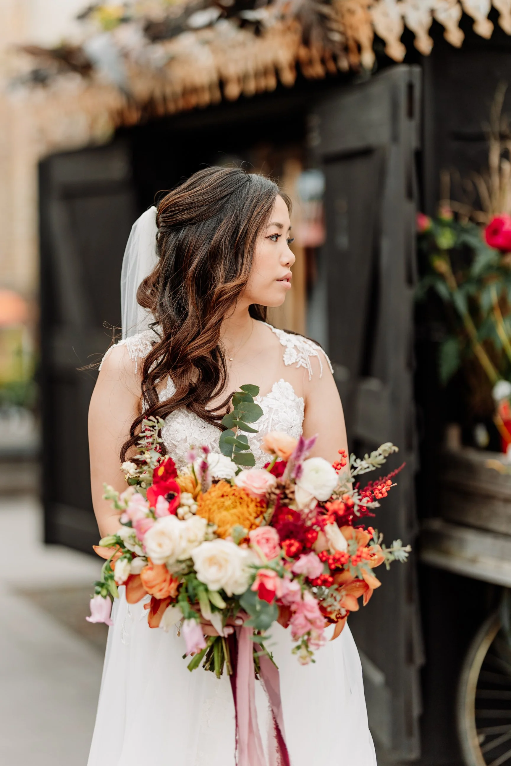 A bride with long wavy dark hair and a white lace wedding dress holding a bouquet of orange, pink, white, and red flowers, standing outdoors.