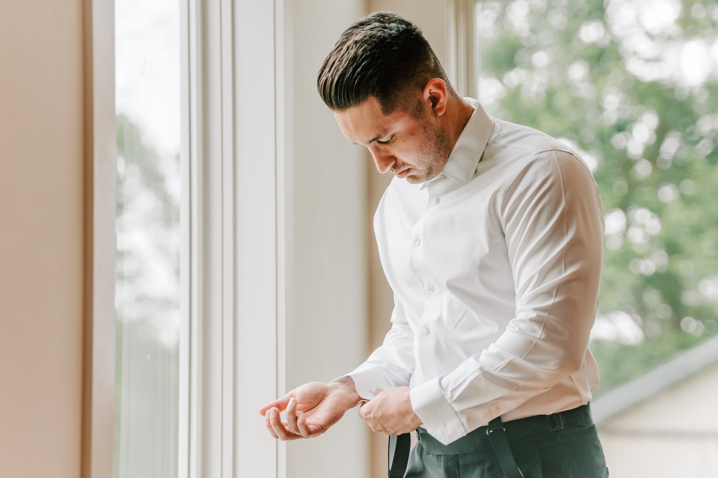 A man in a white dress shirt adjusting his cuff near a window with green trees outside.