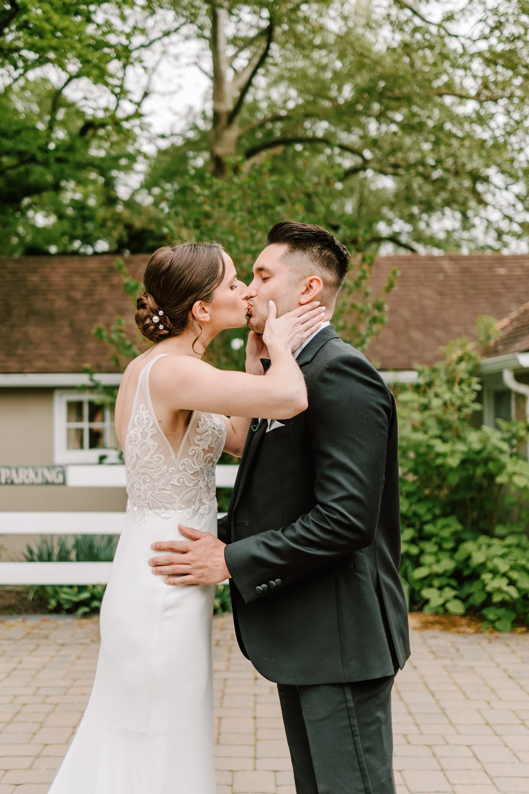 A bride and groom sharing a kiss outdoors on a paved path, with greenery and trees in the background.