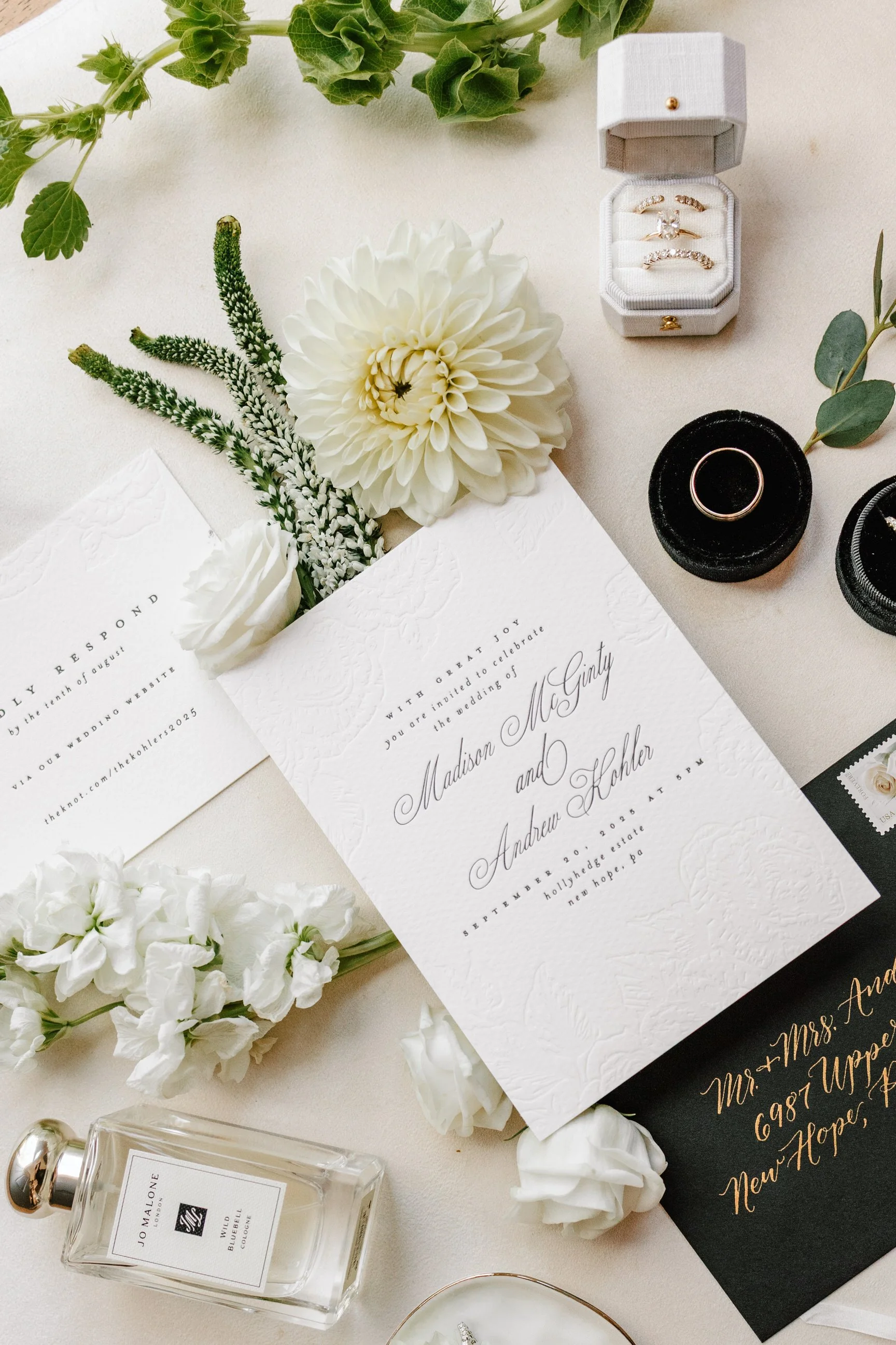 Wedding invitation with white flowers, a jewelry box with rings, a black ring box with a ring, a perfume bottle, and an envelope with gold calligraphy address on a light surface.