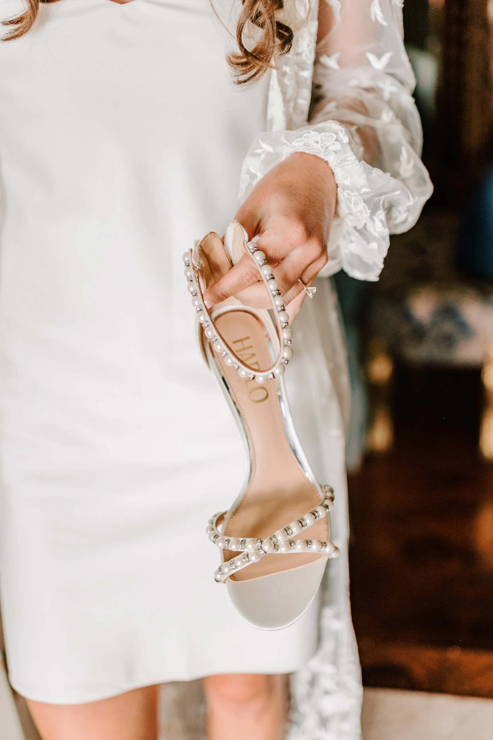 A person in a white dress holding a pearl-embellished beige high heel sandal.