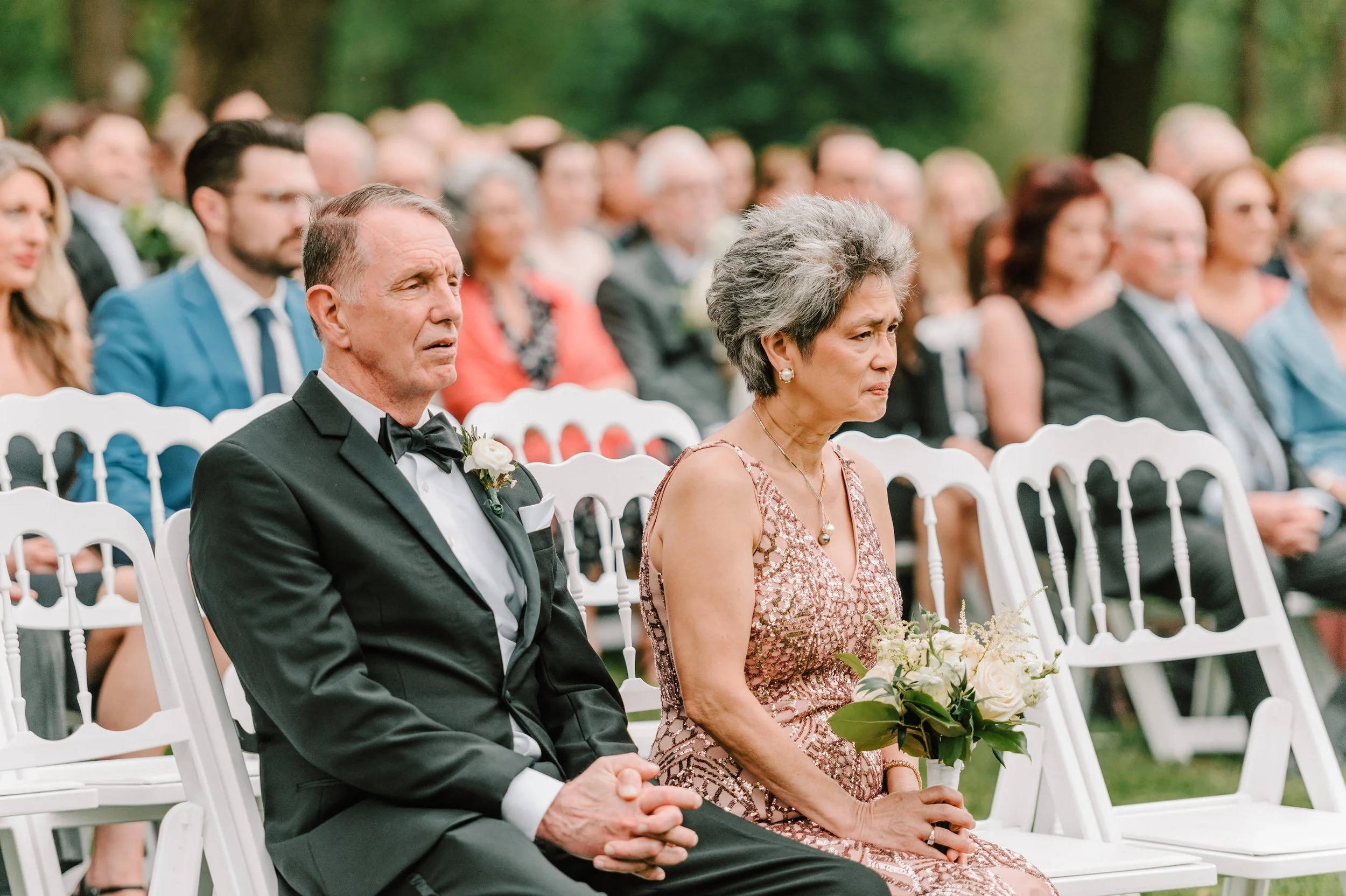 An elderly man and woman sitting in white chairs at an outdoor wedding, with guests seated behind them. The man wears a black tuxedo with a bow tie, and the woman wears a pink lace dress, holding a bouquet of white roses and greenery.