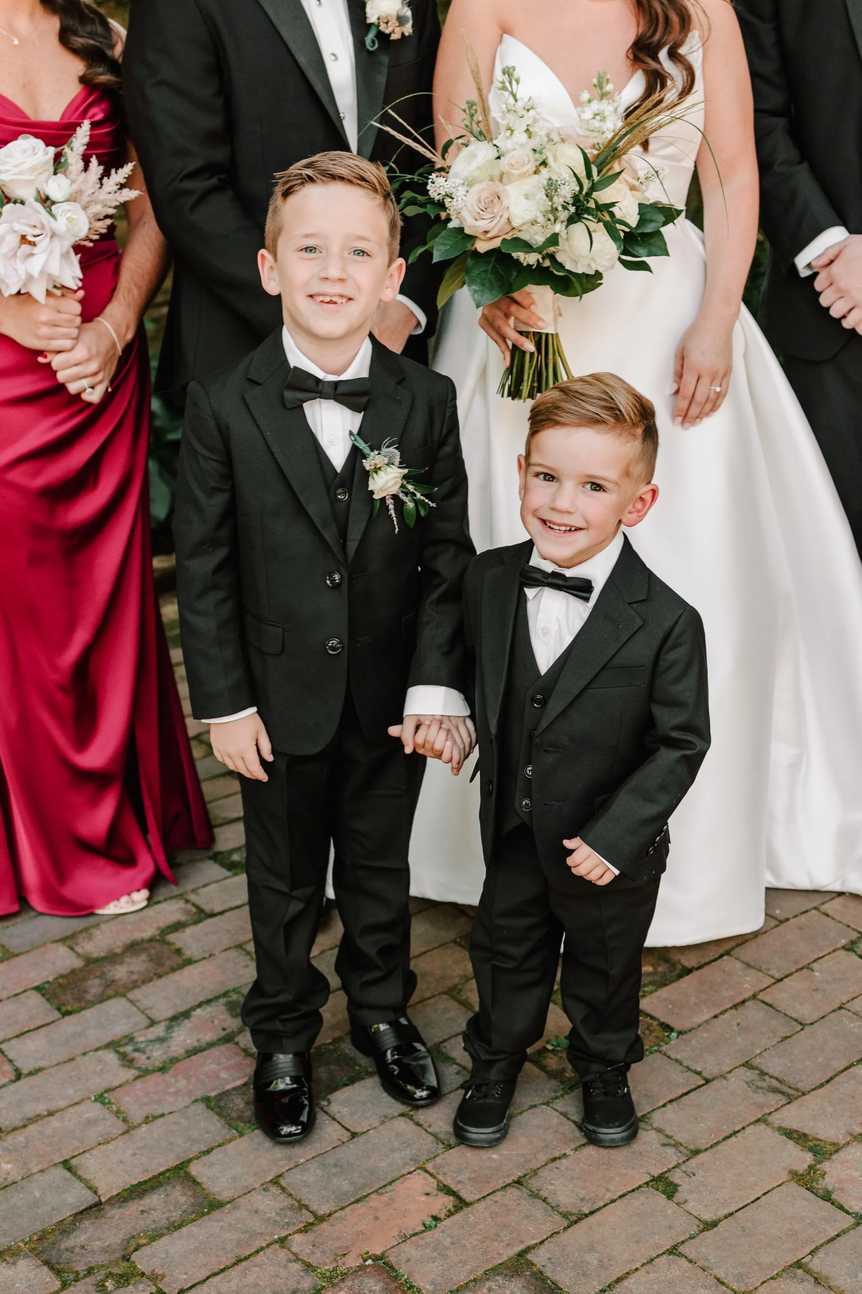 Two young boys in tuxedos holding hands at a wedding, with a bride and a bridesmaid partially visible in the background.