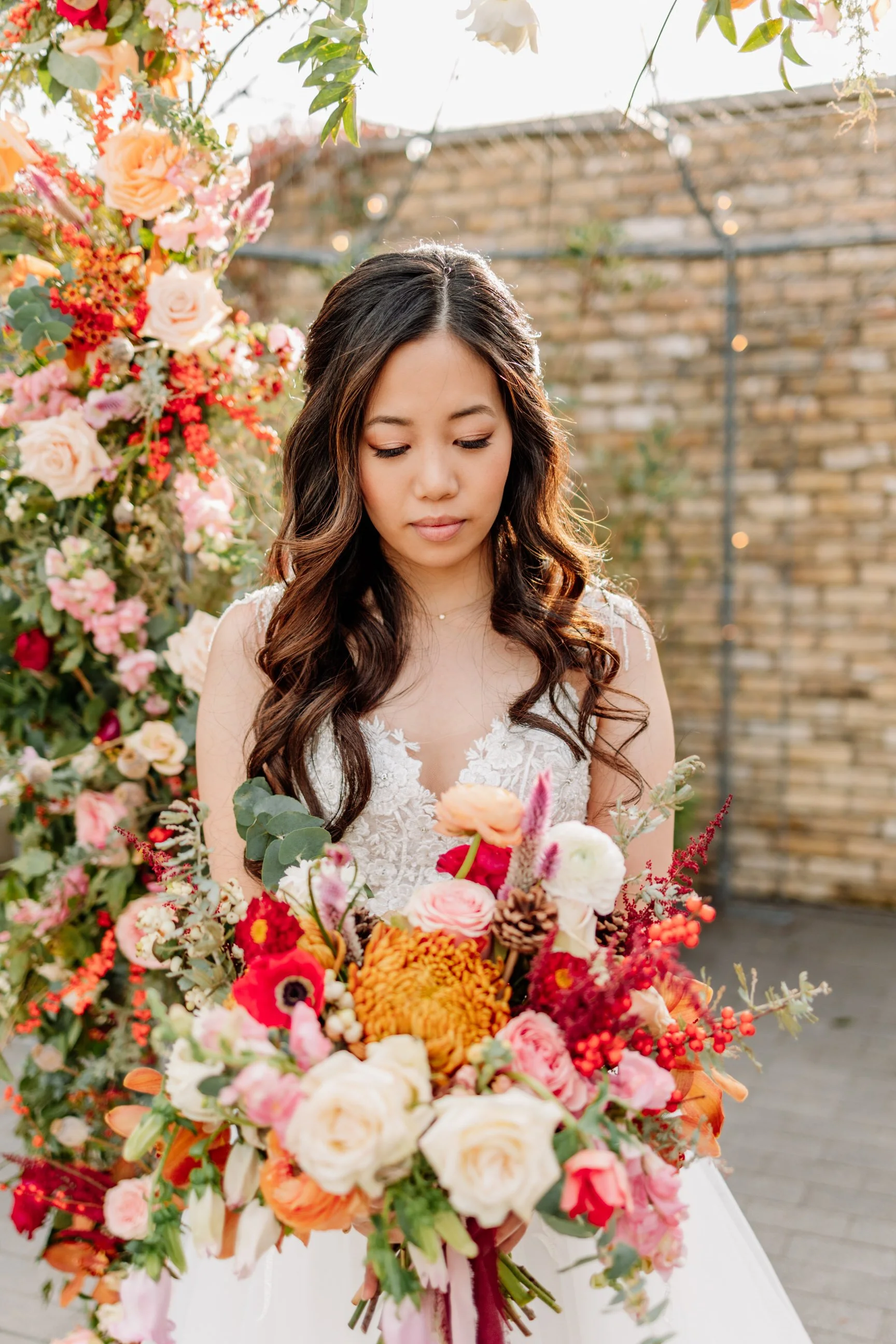 Young woman with long, wavy dark hair in a white lace dress holding a colorful bouquet of flowers, standing outdoors near a flower arrangement and brick wall.