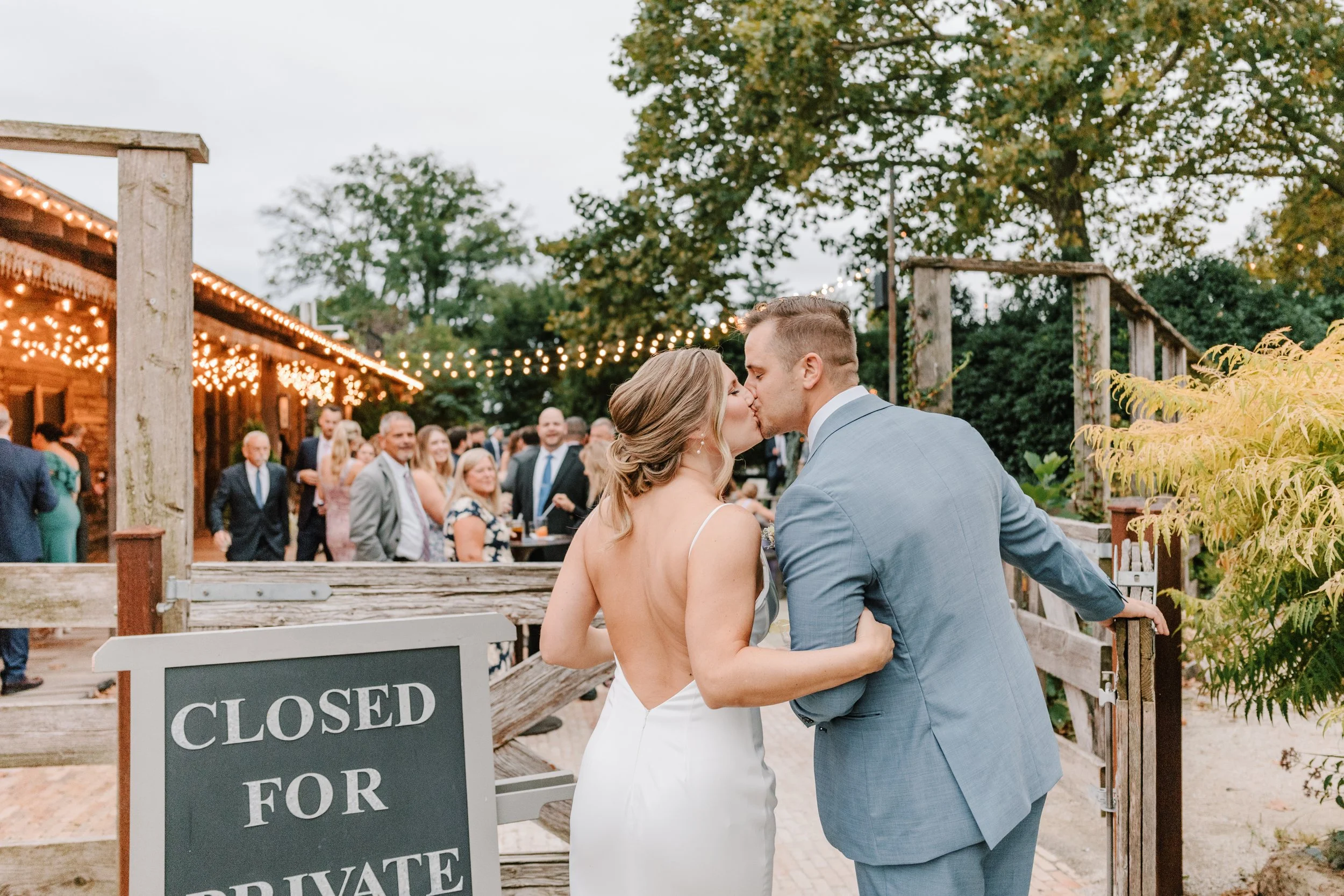 A newlywed couple sharing a kiss at their outdoor wedding reception, with guests in the background and string lights overhead. Philadelphia wedding photographer