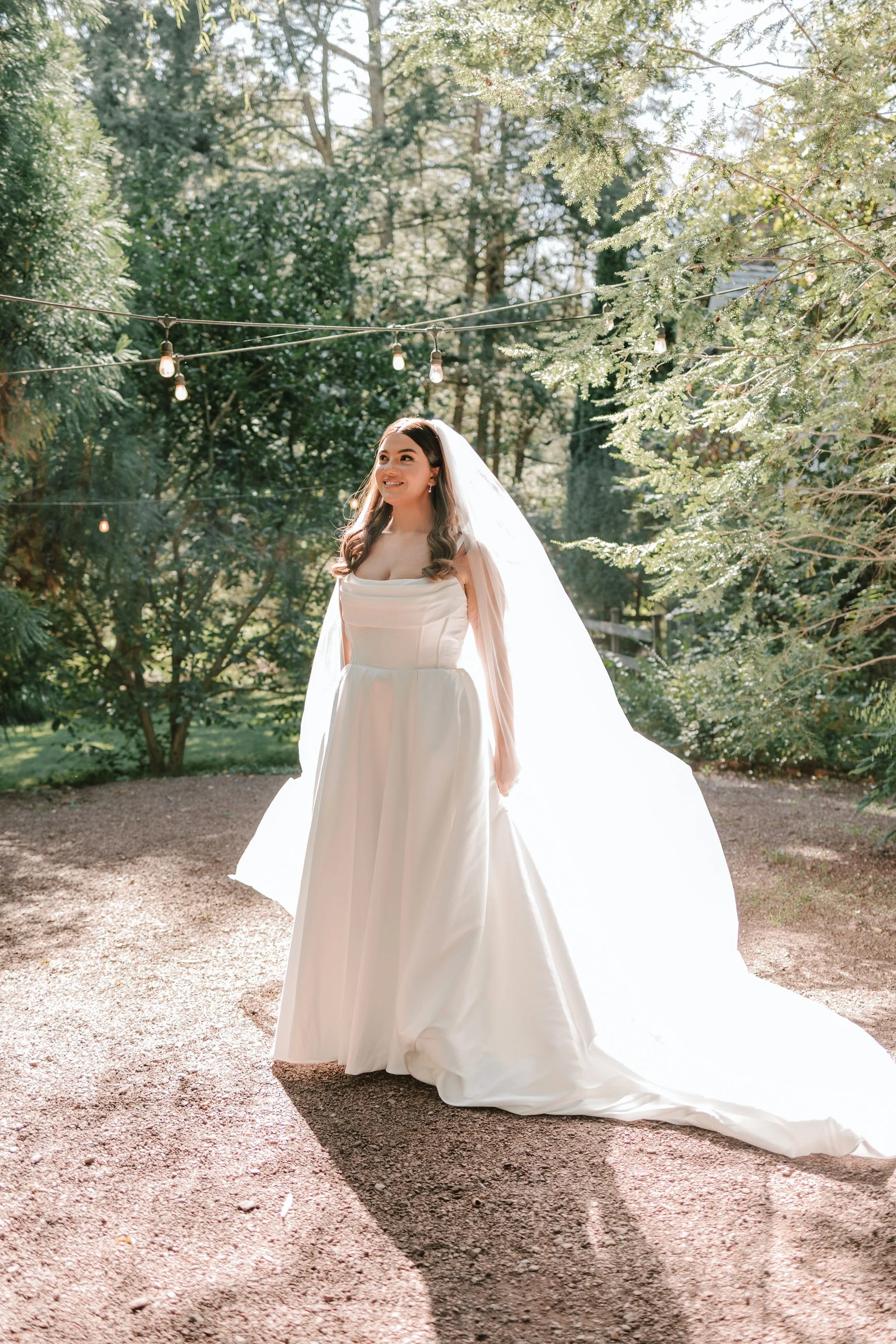 Bride in a white wedding gown standing outdoors on a gravel path with trees and string lights overhead, sunlight filtering through the foliage.