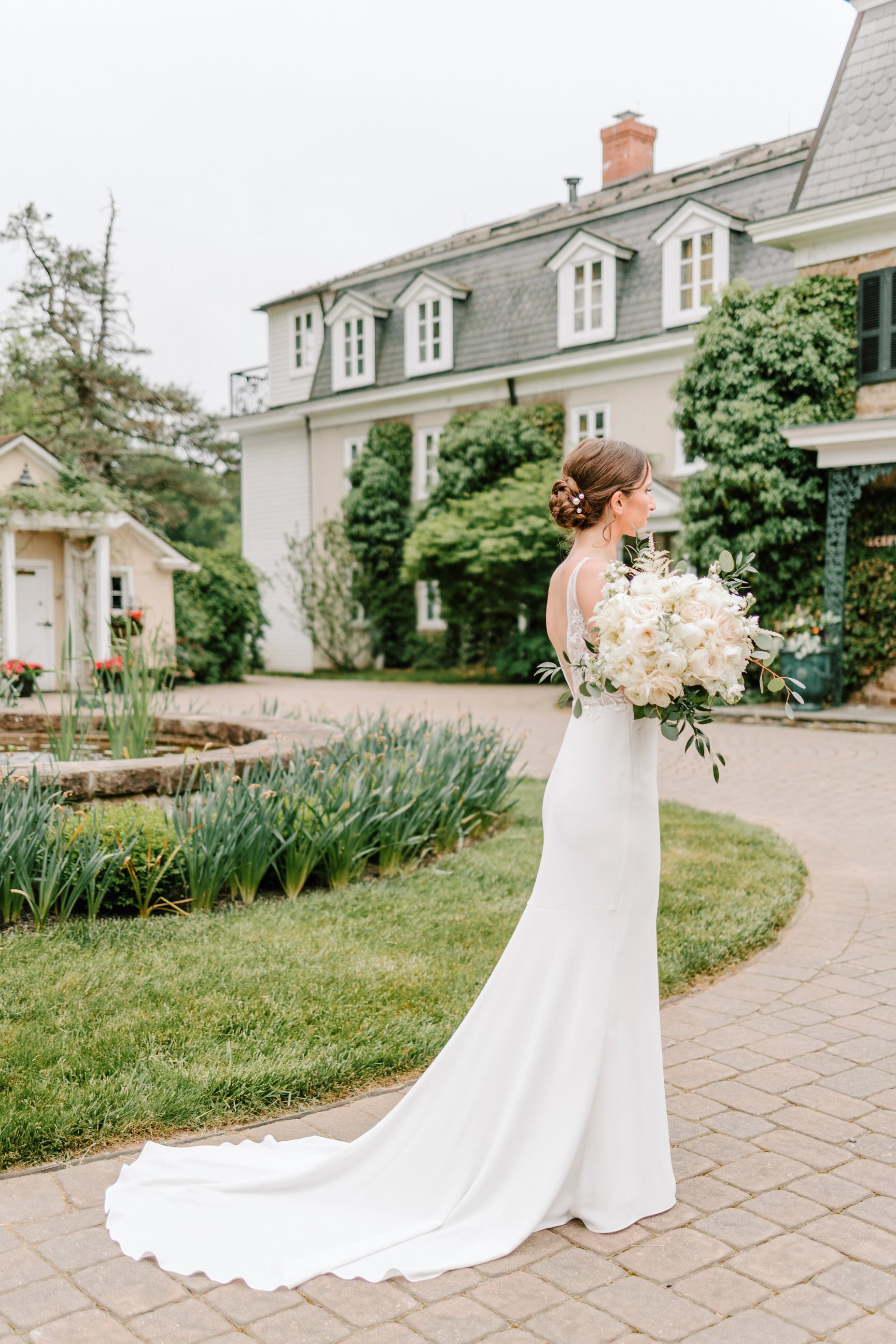 Bride in a white wedding dress holding a large bouquet of white roses and greenery, standing outdoors on a paved path near a garden with green shrubs and flowers, with a large house in the background.