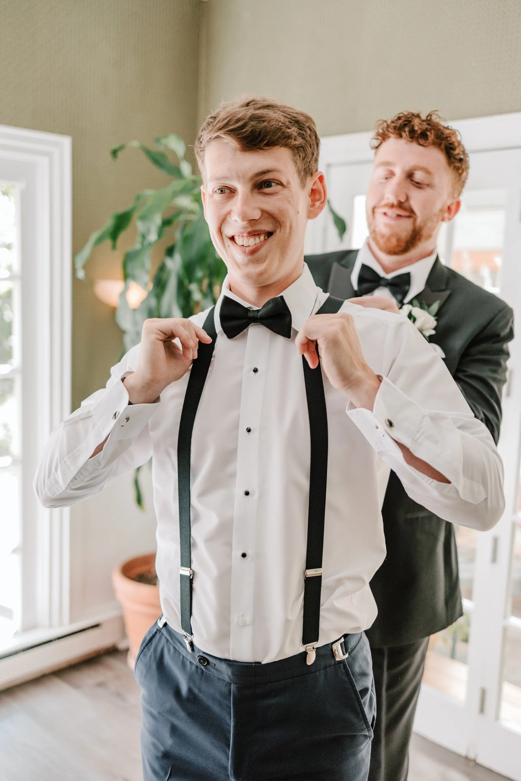 Two men in tuxedos, one with suspenders, preparing for a formal event in a bright room with large windows and potted plants.