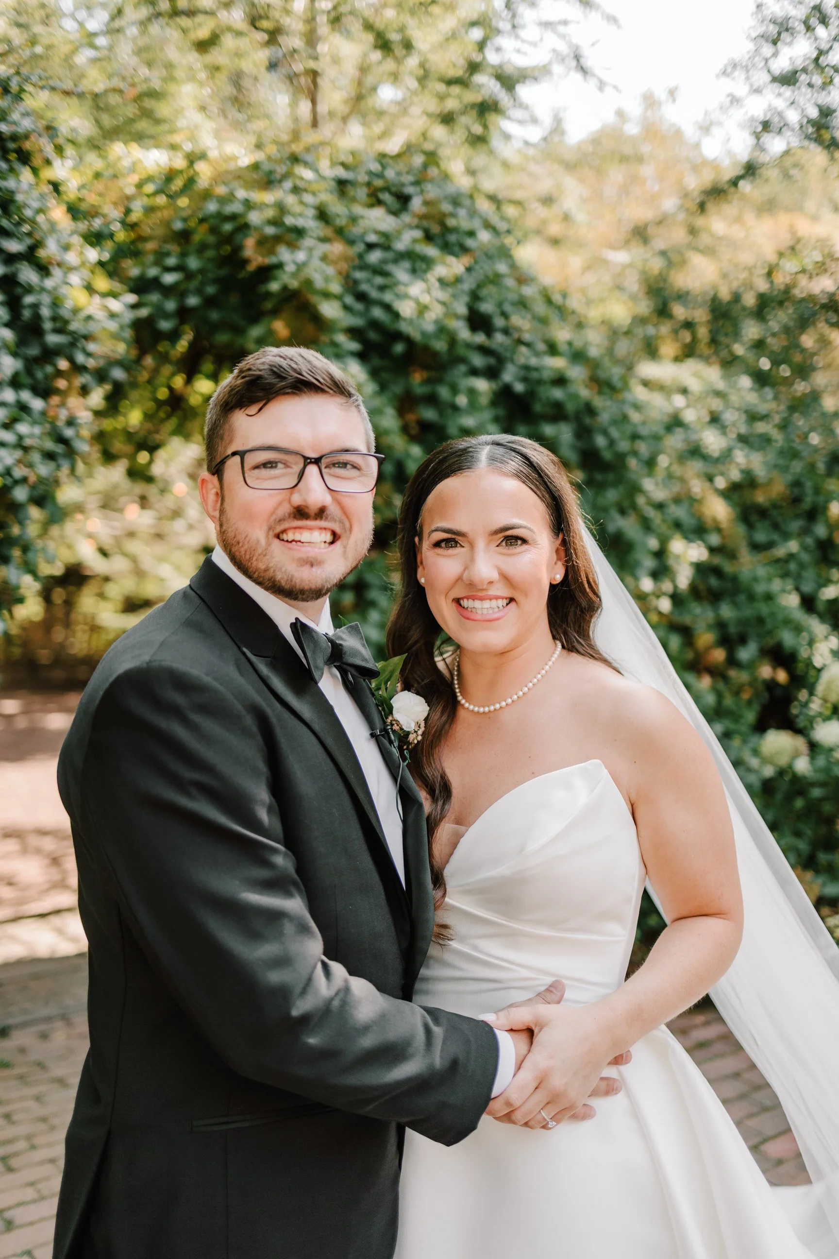 A newlywed couple smiling while holding hands outdoors during daytime, with greenery in the background.