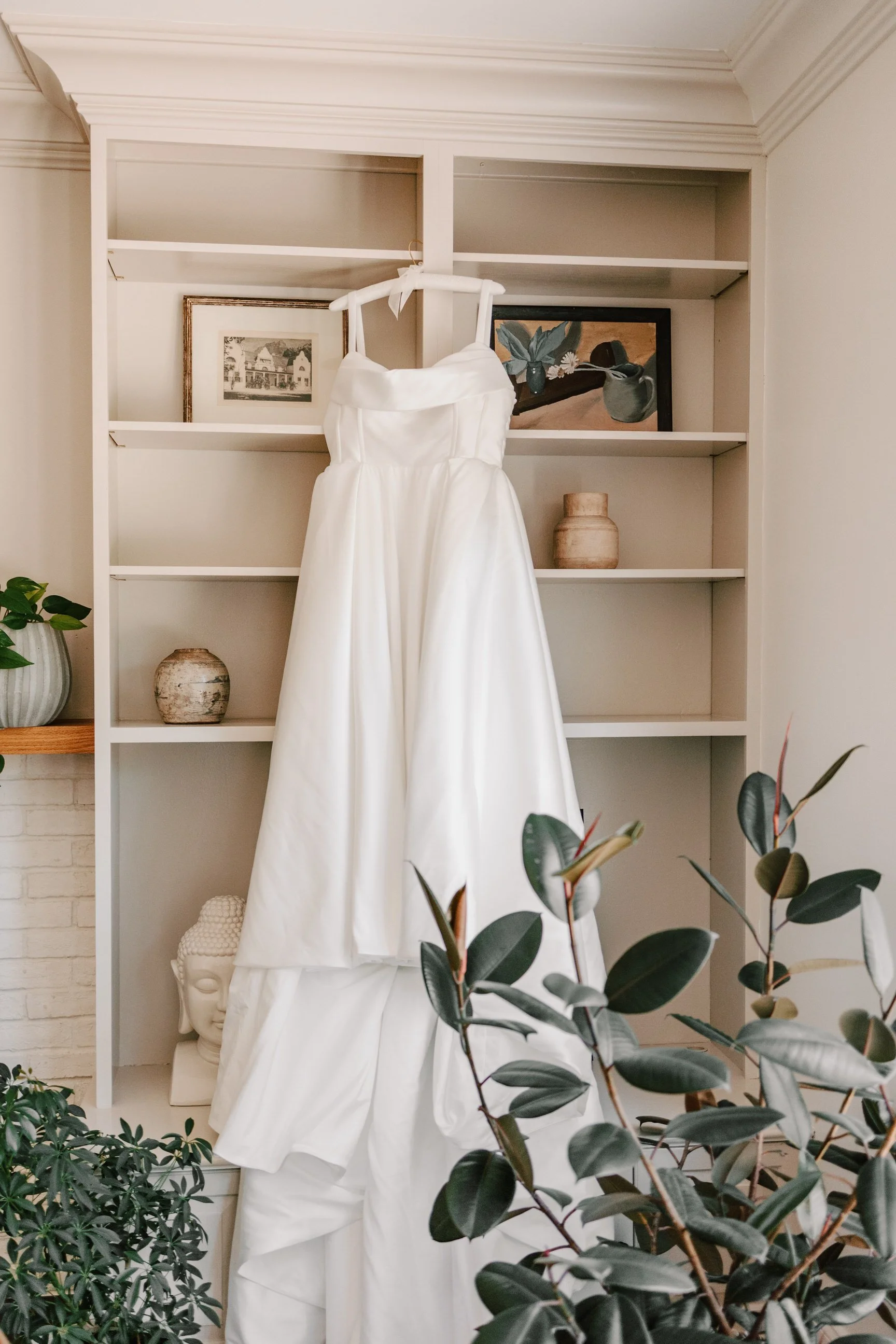 A white wedding dress hanging in front of a bookshelf, surrounded by green plants and decorative items.