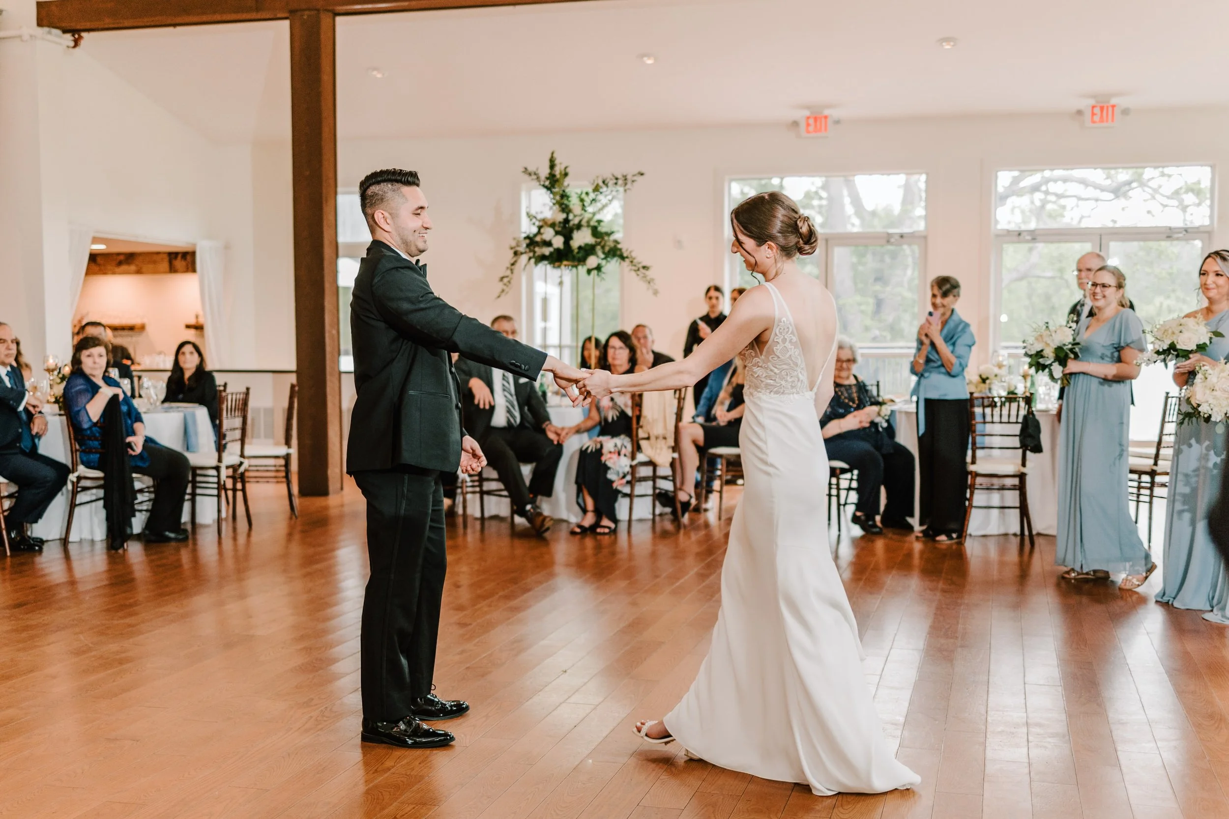  Philadelphia wedding photographerBride and groom holding hands during their wedding dance in a reception hall, with guests seated and standing around them, some taking photos and videos.