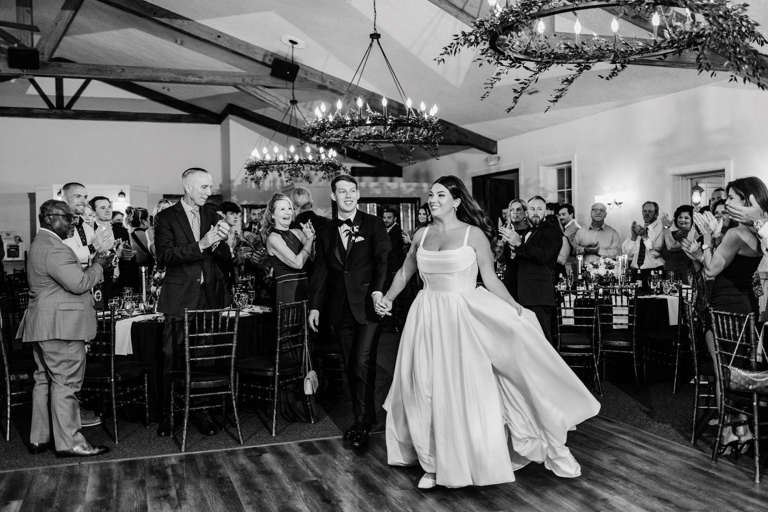 A bride and groom walking hand-in-hand on the dance floor during their wedding reception, surrounded by seated and standing guests clapping and smiling in a decorated indoor venue.