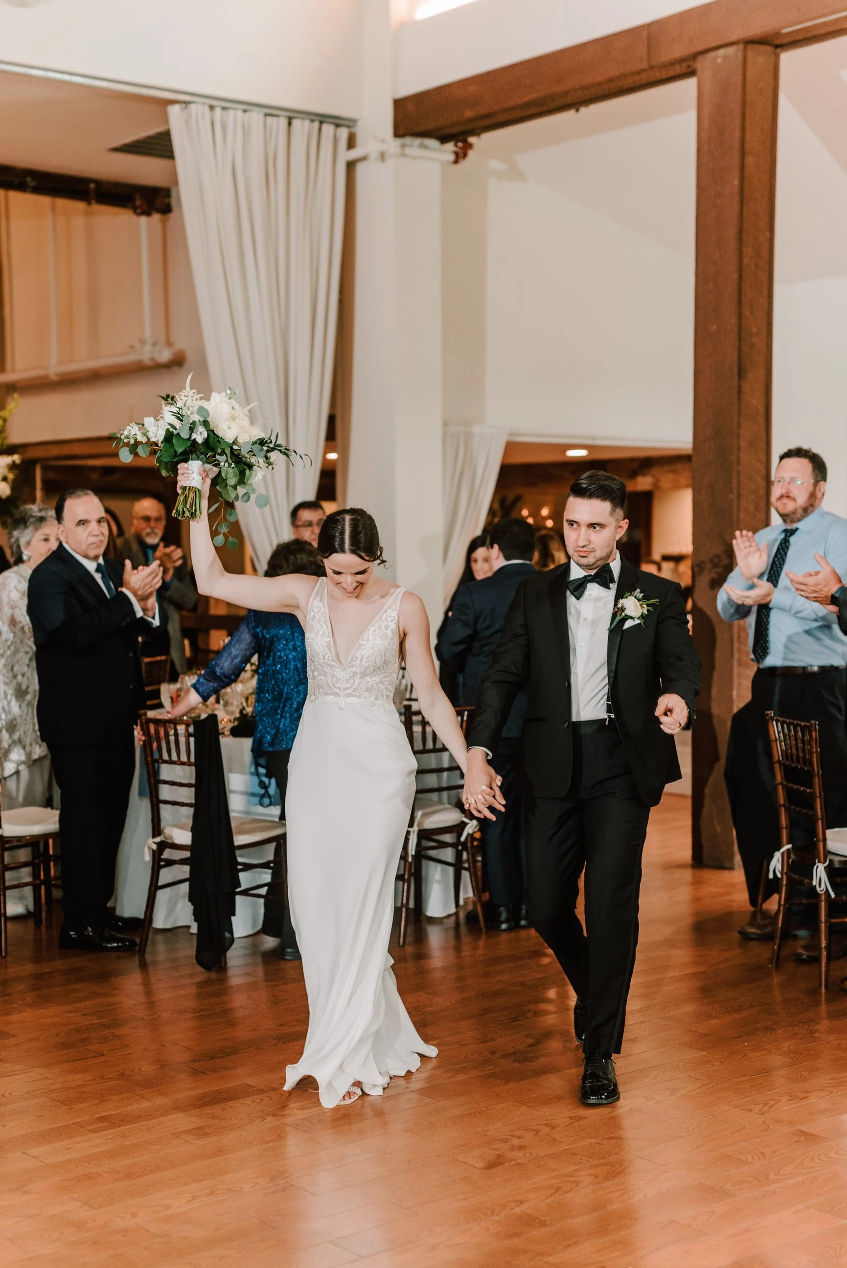 Bride and groom walking hand in hand during their wedding reception, guests applauding in the background.