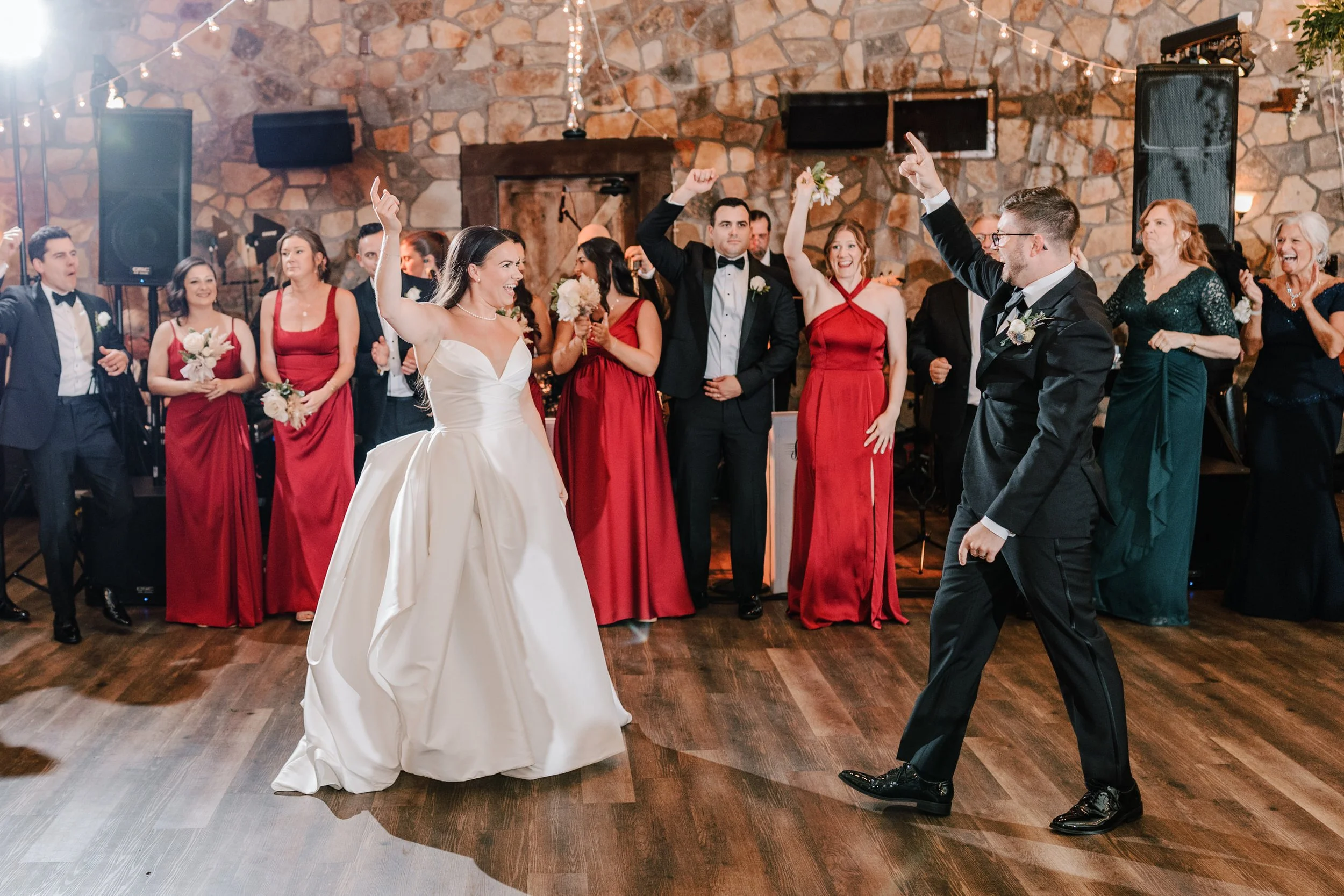 Bride and groom dancing at their wedding reception, surrounded by bridesmaids and groomsmen in formal attire, with a stone wall in the background.