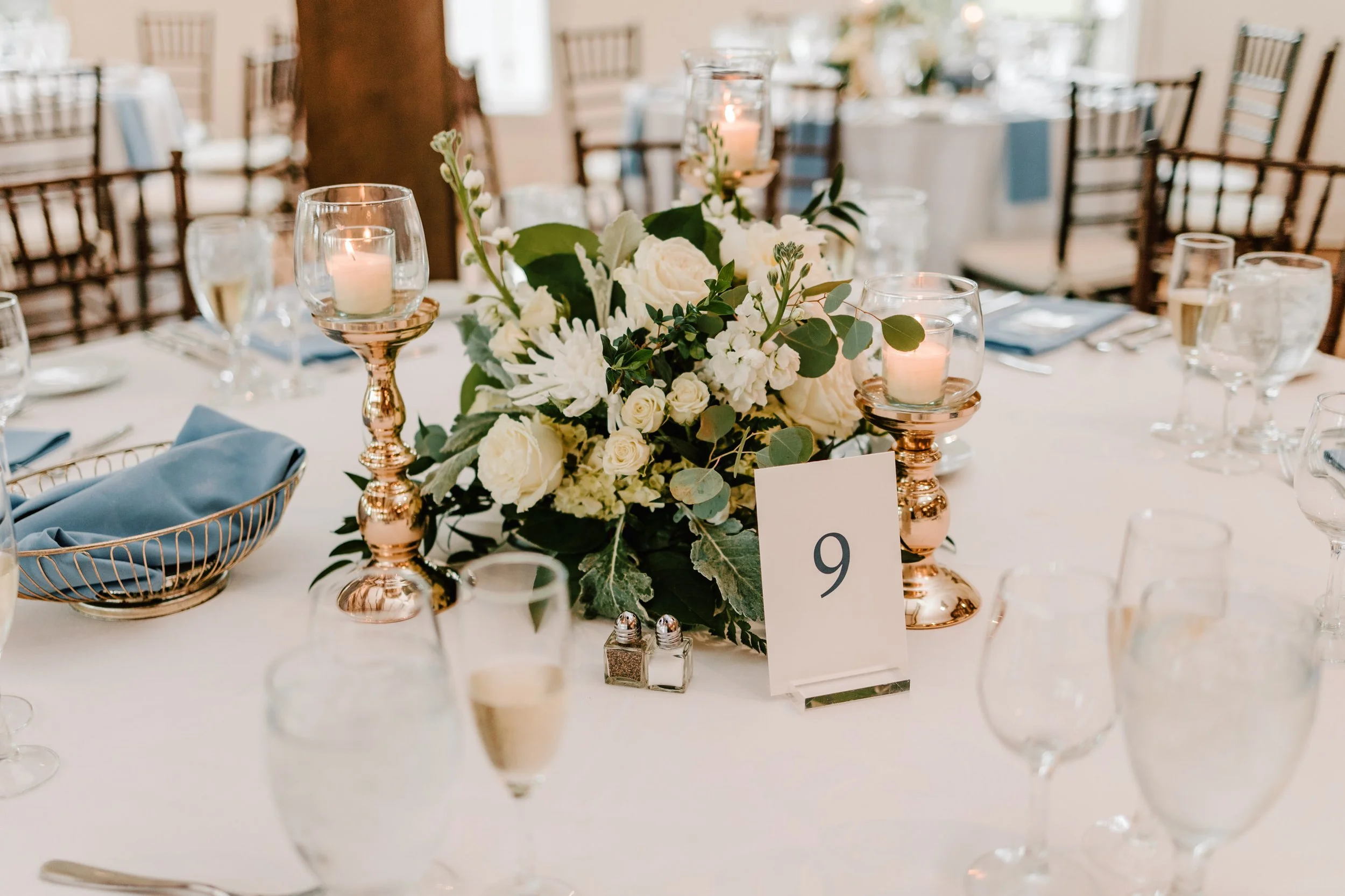 A decorated table at a wedding or formal event with a floral centerpiece, candle holders with lit candles, a table number card with the number 9, champagne glasses, blue napkins, and place settings.