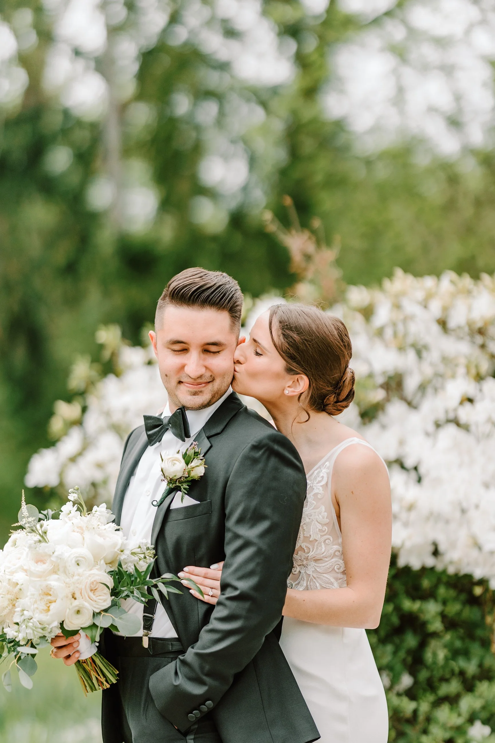 Bride kisses groom's cheek outdoors surrounded by white flowers and greenery.