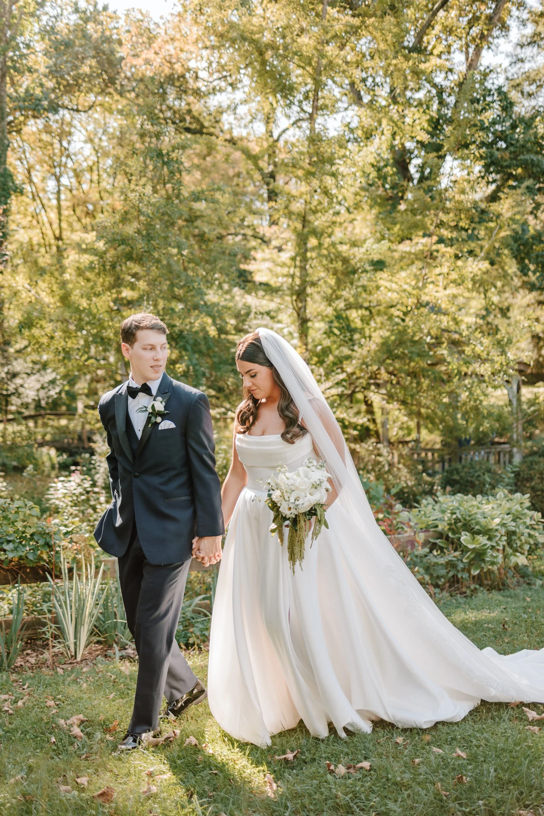 A bride and groom holding hands outdoors in a garden during sunset, surrounded by greenery and trees.
