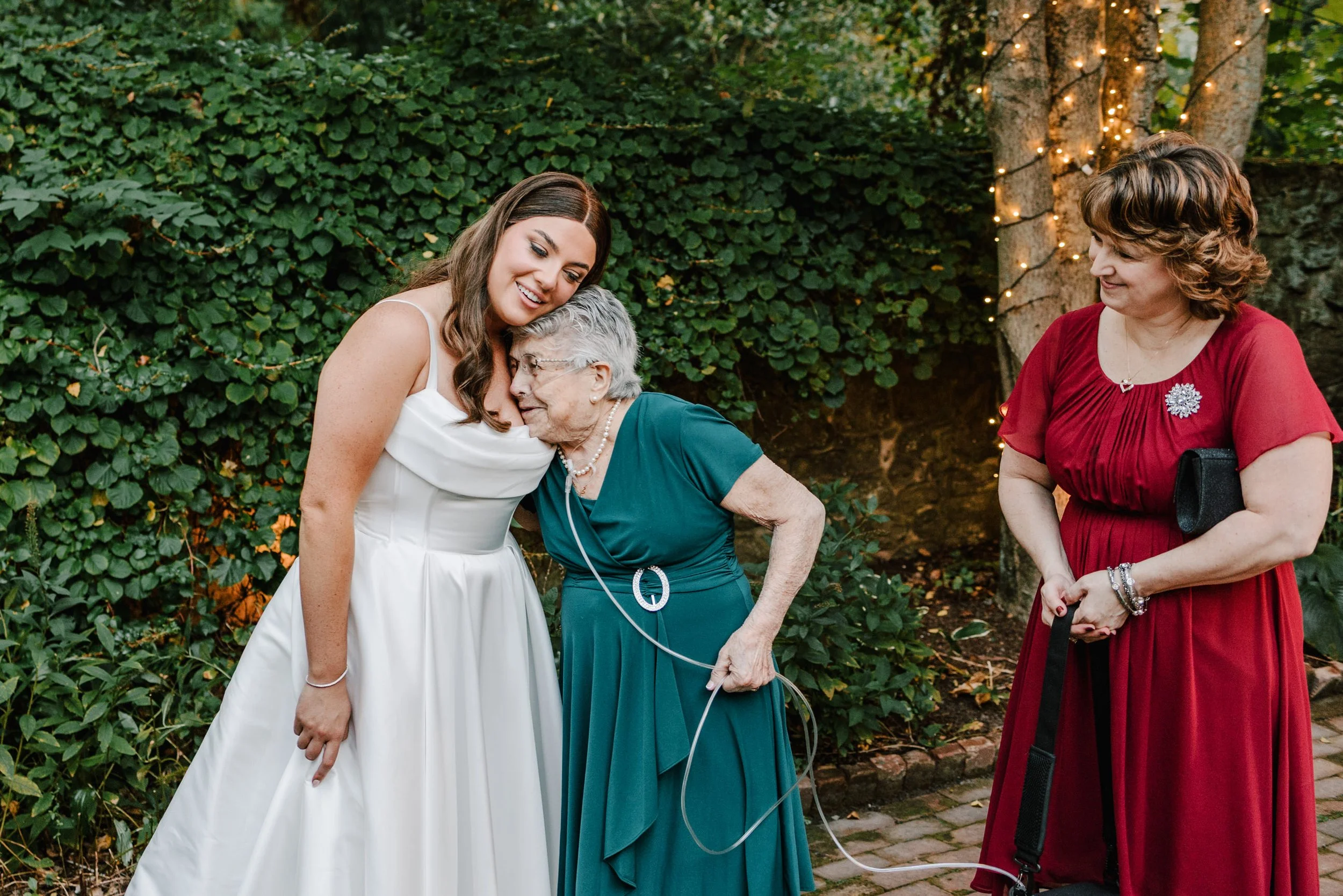 A bride in a white wedding dress is hugging an elderly woman in a teal dress. Two other women, one in a red dress, are standing nearby outdoors among green foliage and string lights.