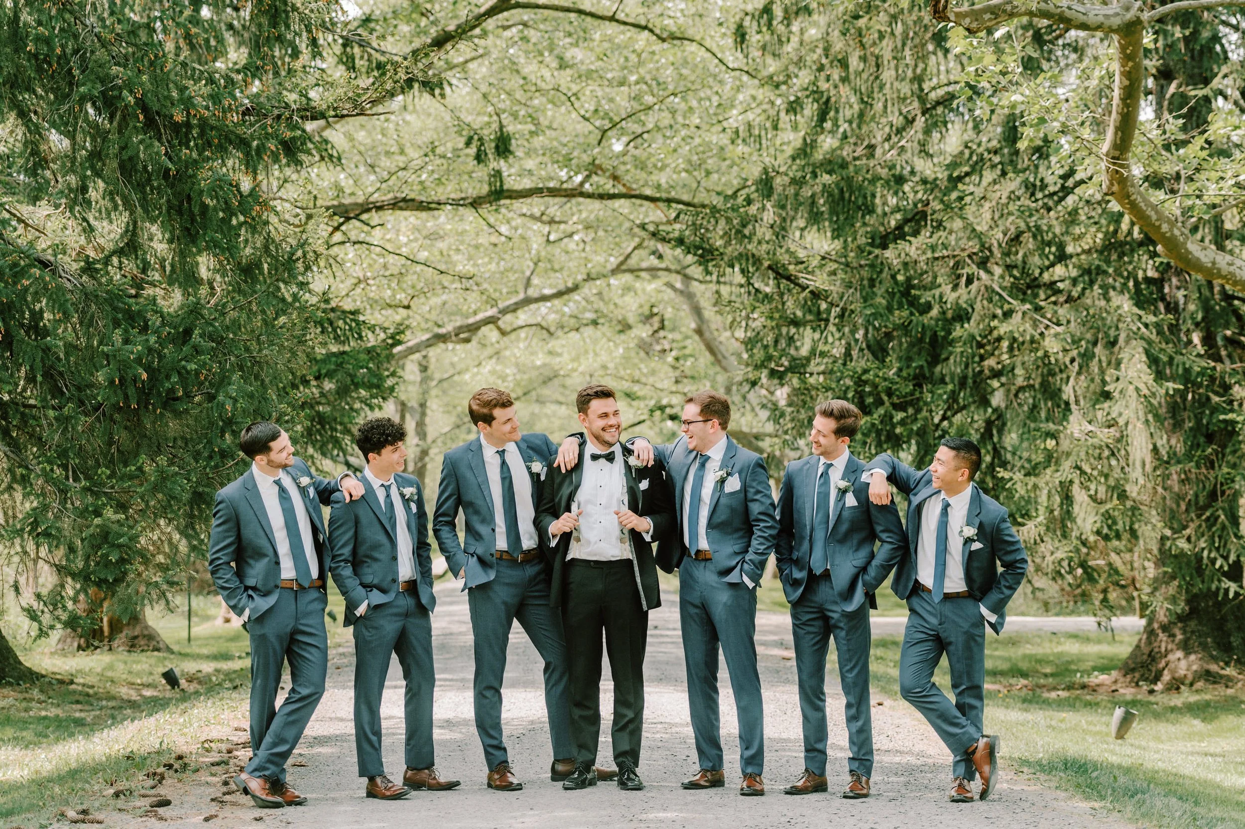 Group of seven men in suits standing outdoors on a path surrounded by trees, smiling and chatting.