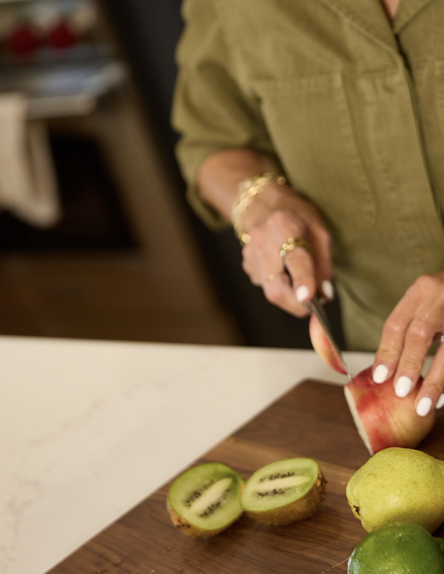Close-up of a Kim Shapira's hands slicing a red and yellow apple on a cutting board, with sliced kiwis and pears nearby.