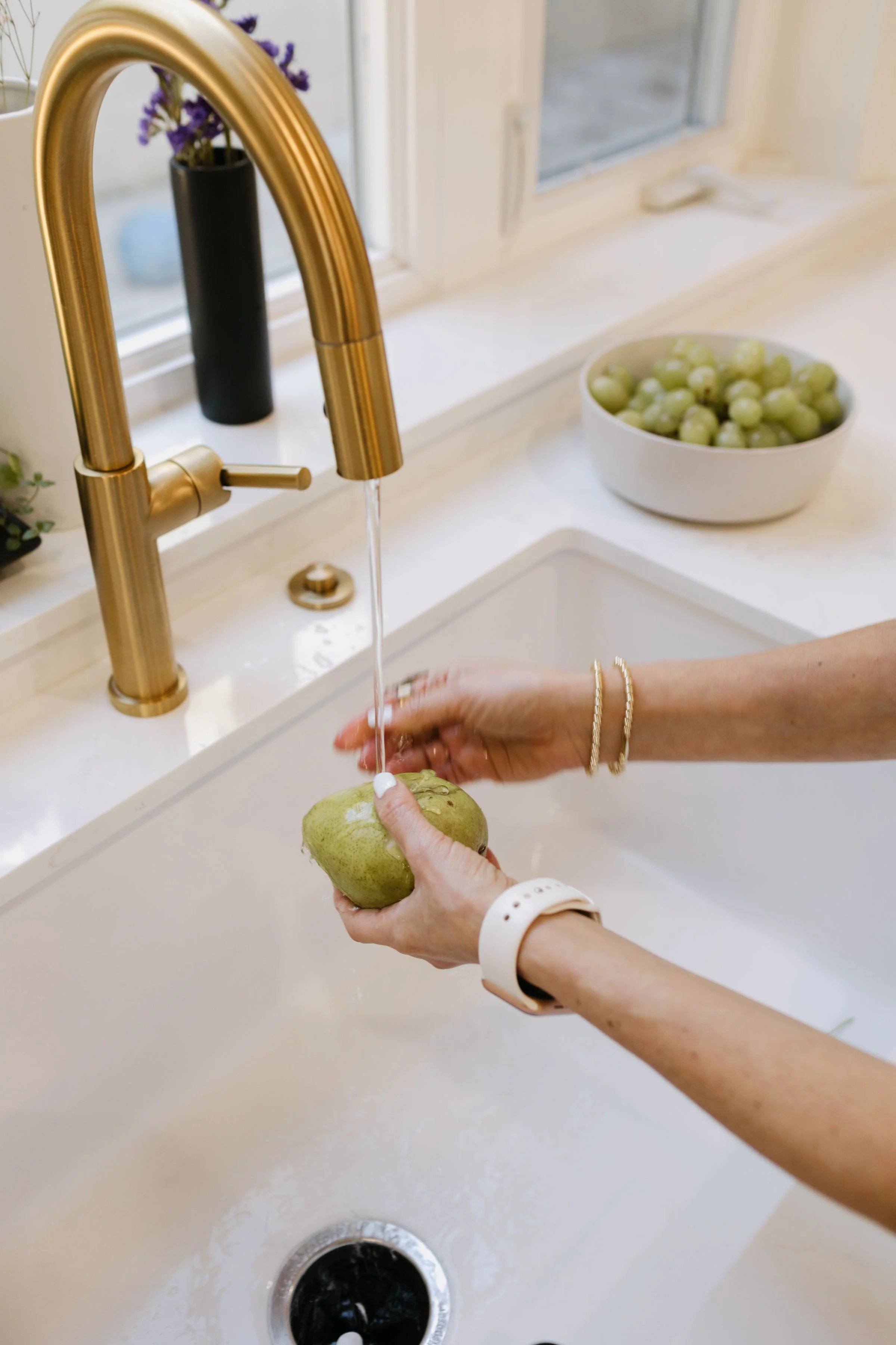 Person washing a pear in a kitchen sink with a gold faucet, with a bowl of green grapes on the countertop behind.