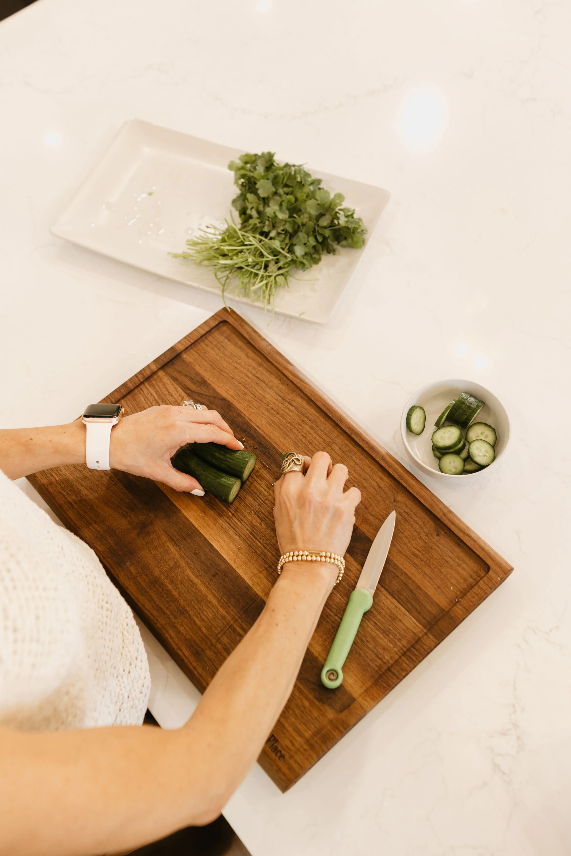 Person chopping cucumber on a wooden cutting board, with sliced cucumber in a bowl nearby and fresh herbs on a plate in the background.