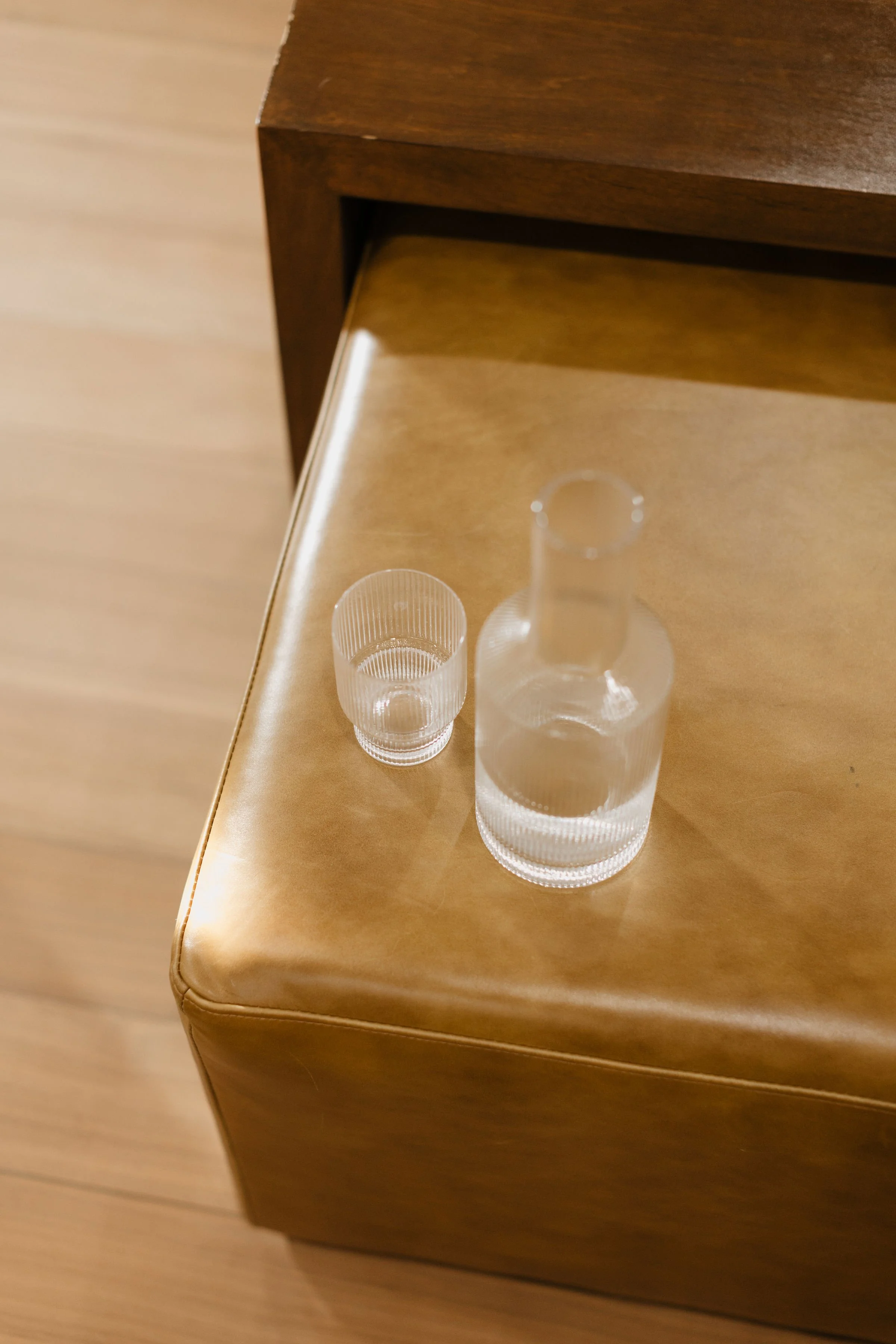 A clear glass bottle and a small clear glass drinking cup on a wooden surface, with a piece of furniture in the background.