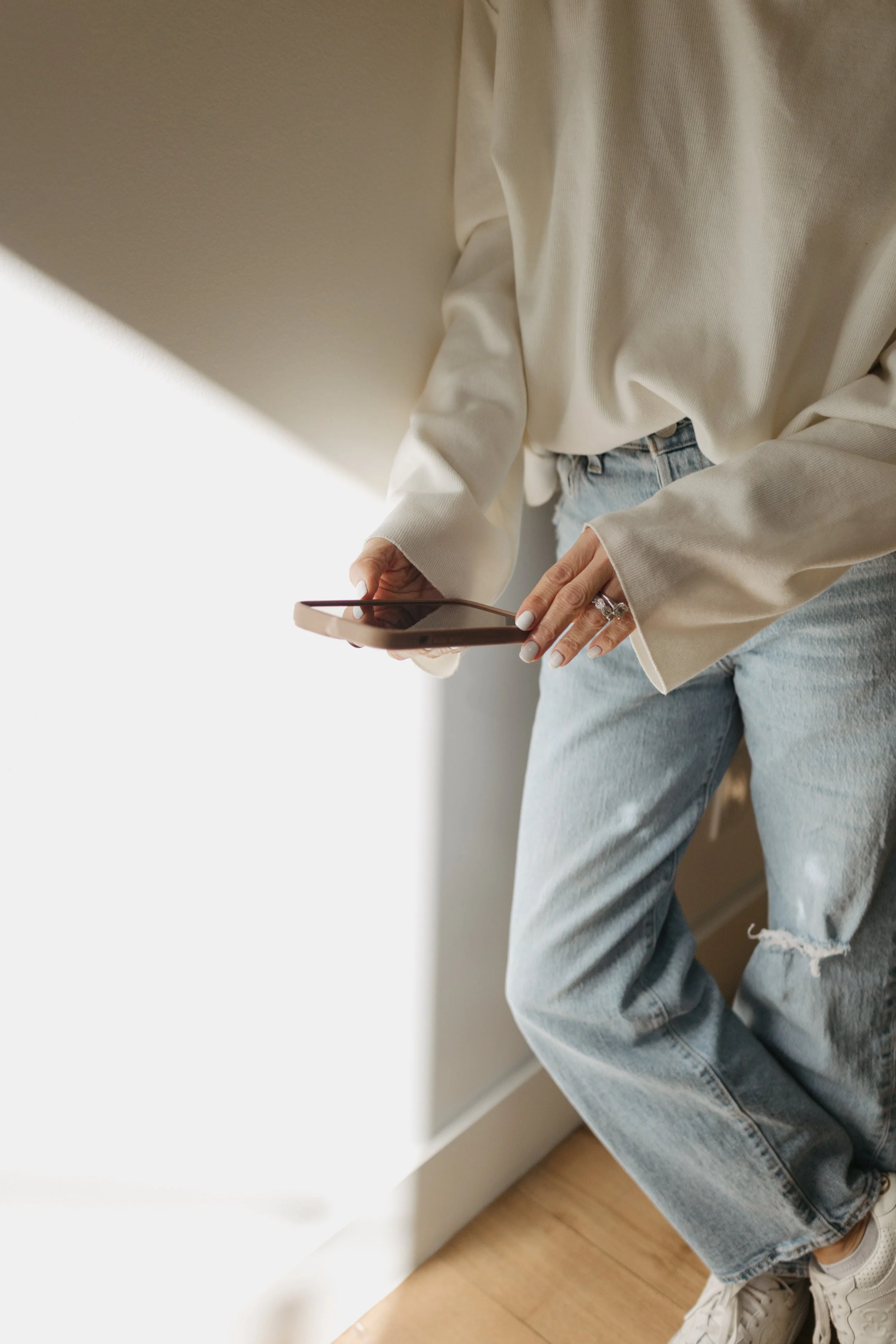 A person dressed in a beige sweater and light blue ripped jeans, holding a smartphone in their right hand, standing near a white wall with sunlight casting a shadow.