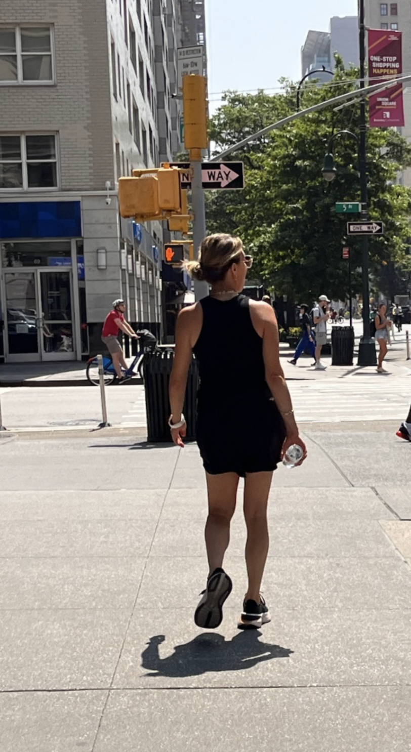 A candid, rear-view photo of Kim Shapira walking down a sunny New York City sidewalk in a black sleeveless top, athletic shorts, and sneakers, carrying a bottle of water.