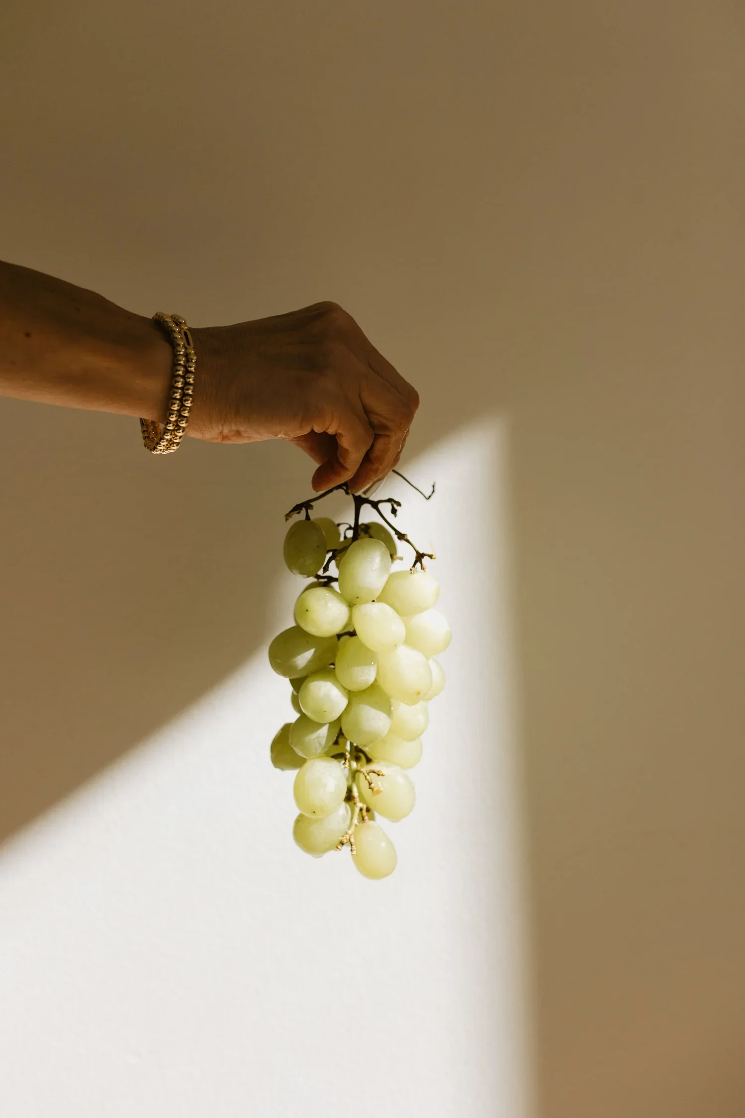 A hand holding a bunch of white grapes against a plain wall with a shadow cast by the grapes.
